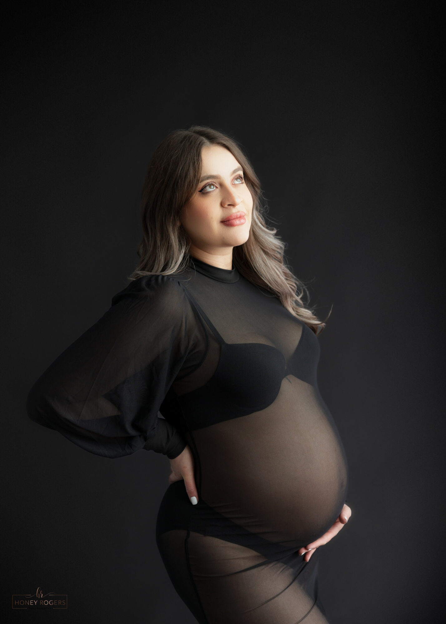 Pregnant woman with long hair in black sheer top, standing against dark background, looking to the side.