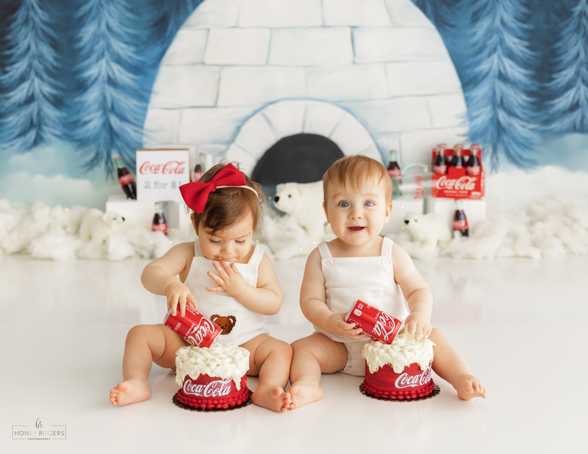 Two children sitting in front of an igloo with Coca-Cola themed decorations, holding whipped cream and wearing red bows and shoes.