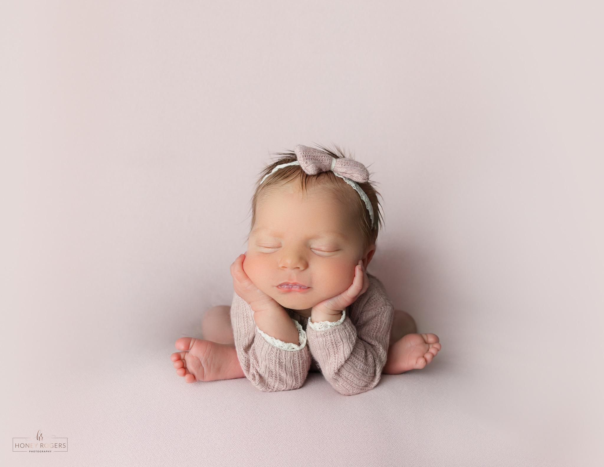 Baby with eyes closed, resting head on hands, wearing a headband and sweater, lying on a light background.