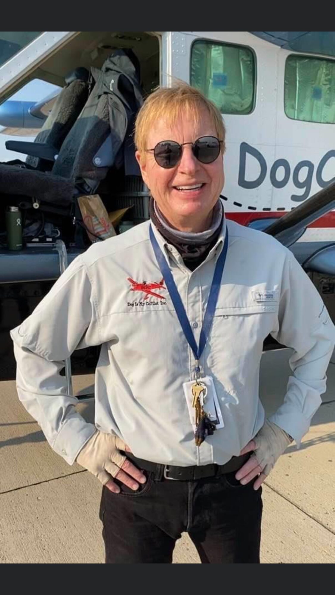 Person with short blonde hair wearing sunglasses, a white jacket, and a lanyard, standing outdoors in front of a helicopter.