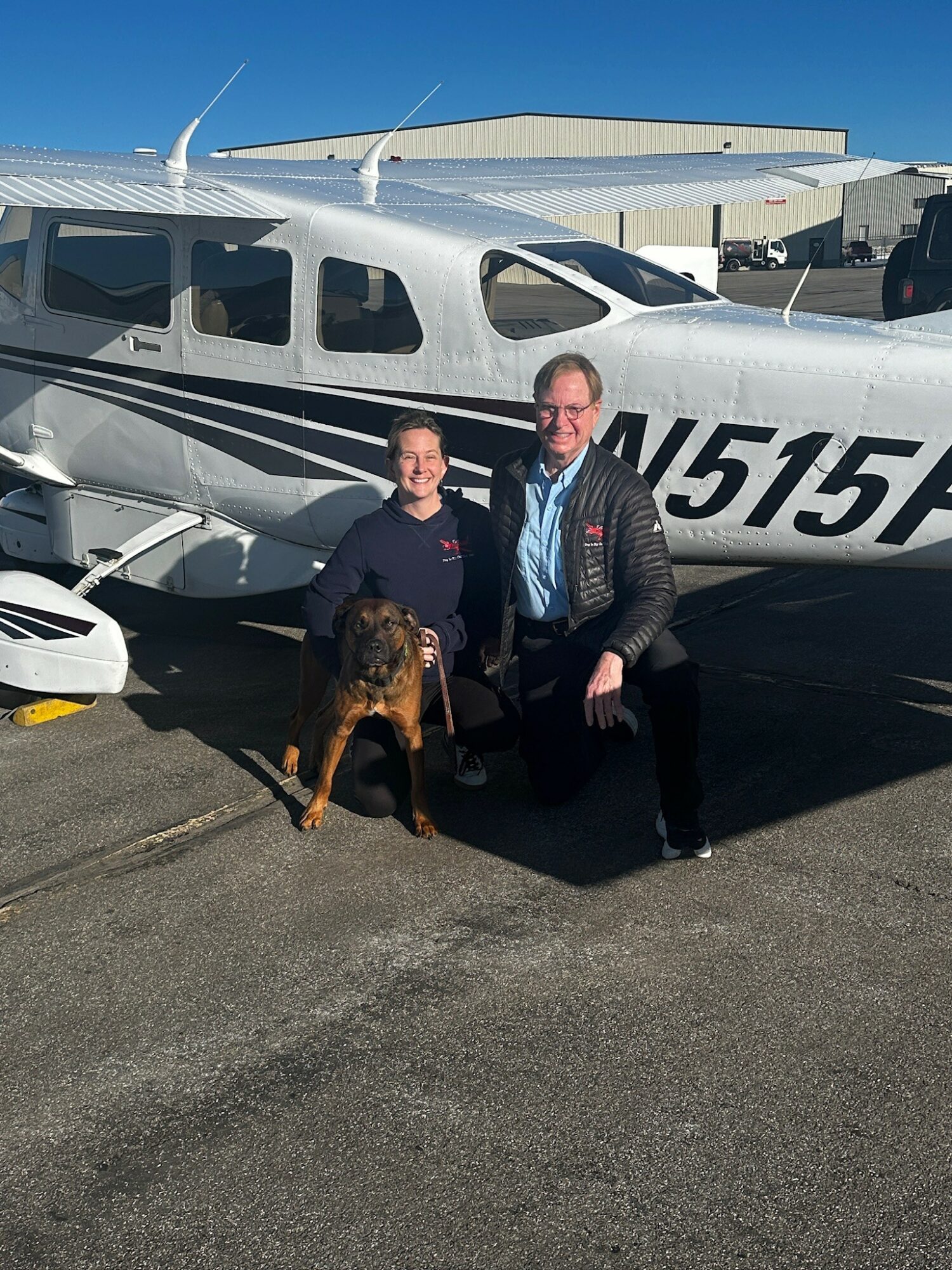 Two people and a dog in front of a small airplane at an airport, clear blue sky in background.