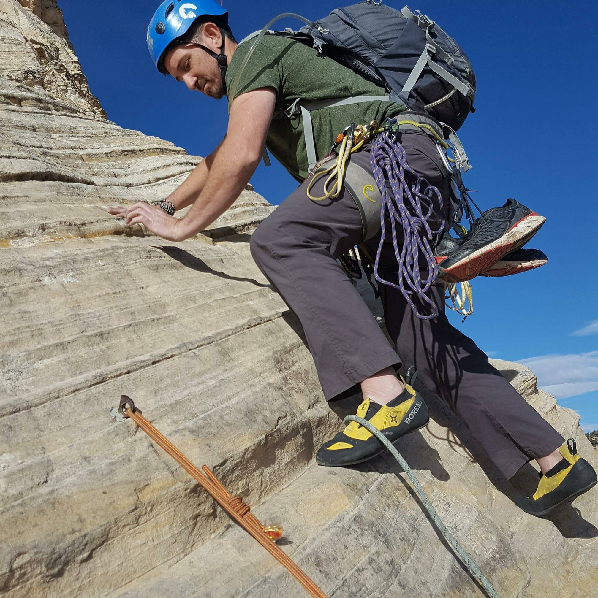 Climber wearing a helmet and backpack ascending a rock face with climbing gear attached, against a blue sky.