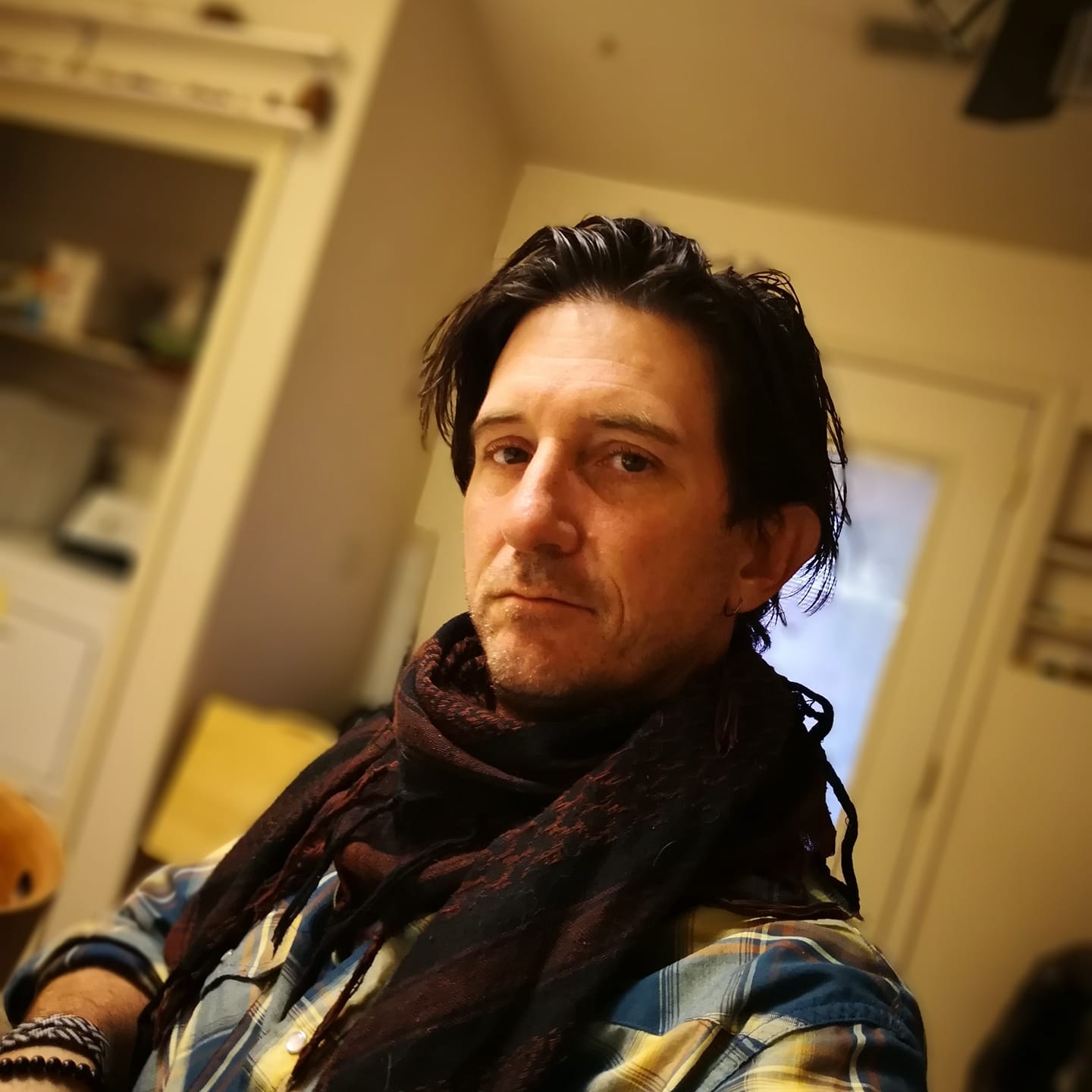 Man with dark hair wearing a plaid shirt and scarf, standing indoors with a neutral expression.
