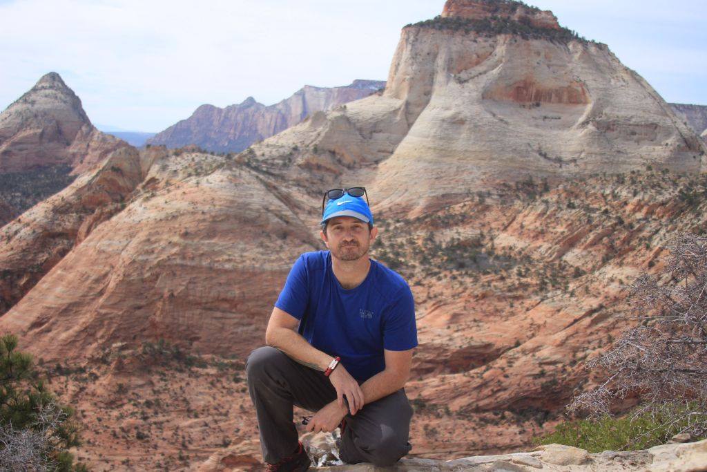 Man sitting on a rock in a canyon with layered rock formations in the background.
