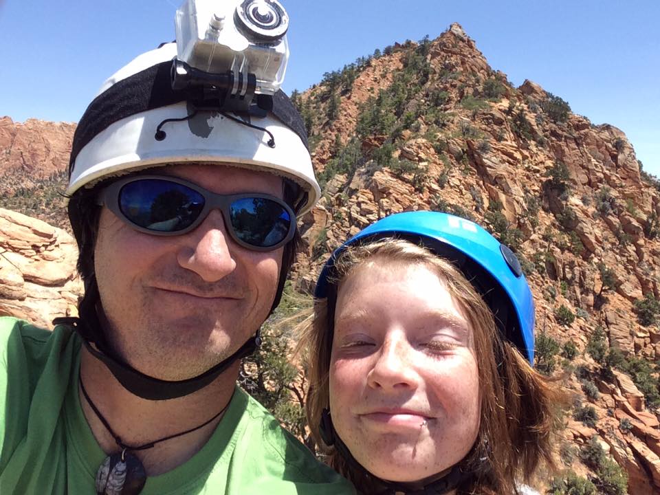 Two people wearing helmets and sunglasses outdoors with rocky mountains in the background.