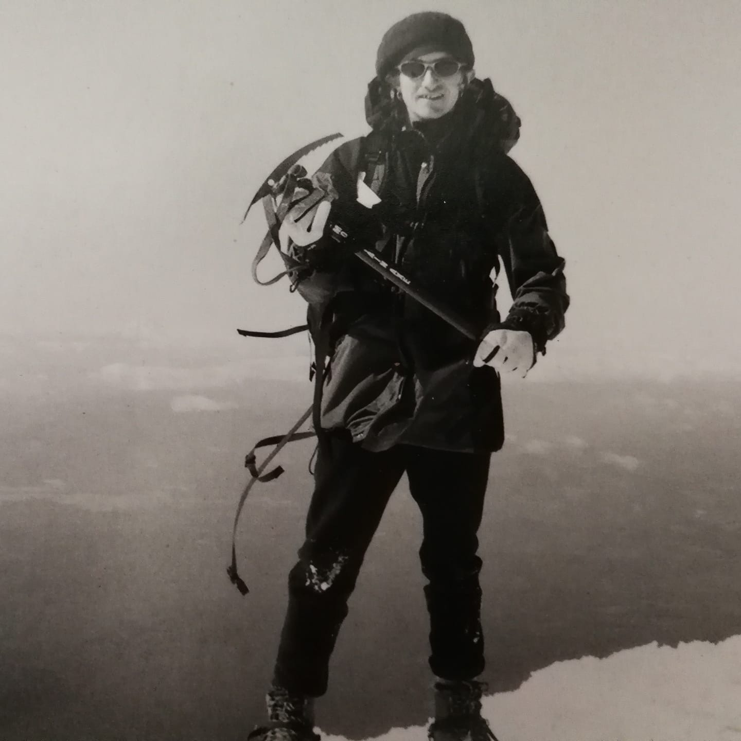 Person standing outdoors in winter clothing, holding an ice axe, with snow and fog in the background.