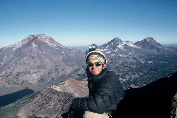 Person wearing sunglasses and a winter hat, standing outdoors with mountains in the background.