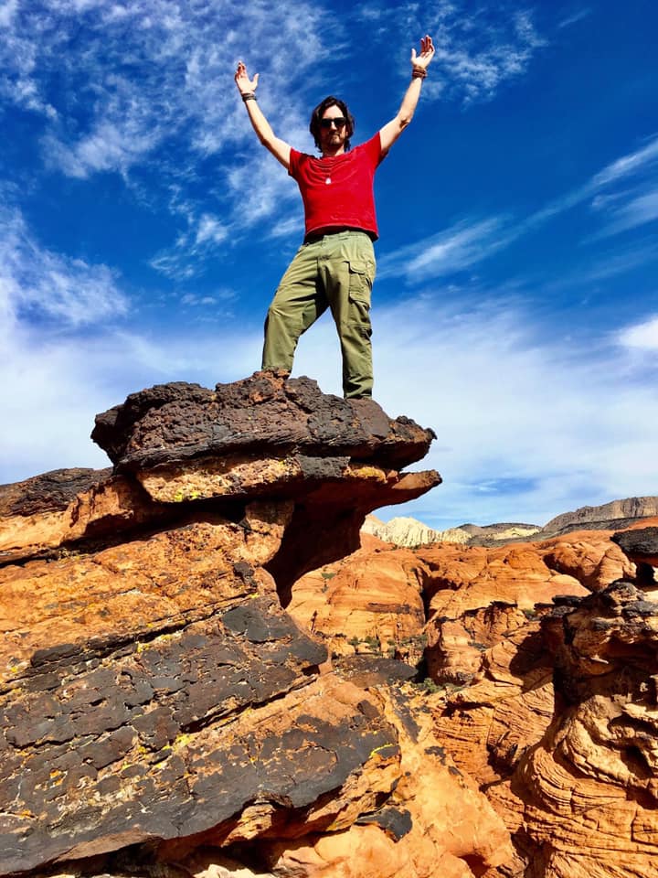 Person standing on rocks with arms raised against a blue sky with clouds, wearing sunglasses, red shirt, and green pants.