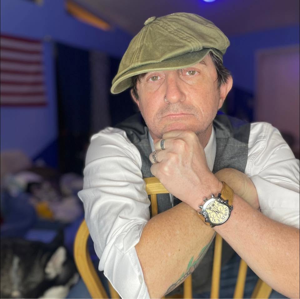 Man wearing a green cap and white shirt, resting his chin on his hand, in a room with blue lighting.