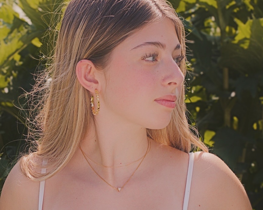 Young woman with long hair, wearing earrings and a necklace, looking to the side outdoors with greenery in background.