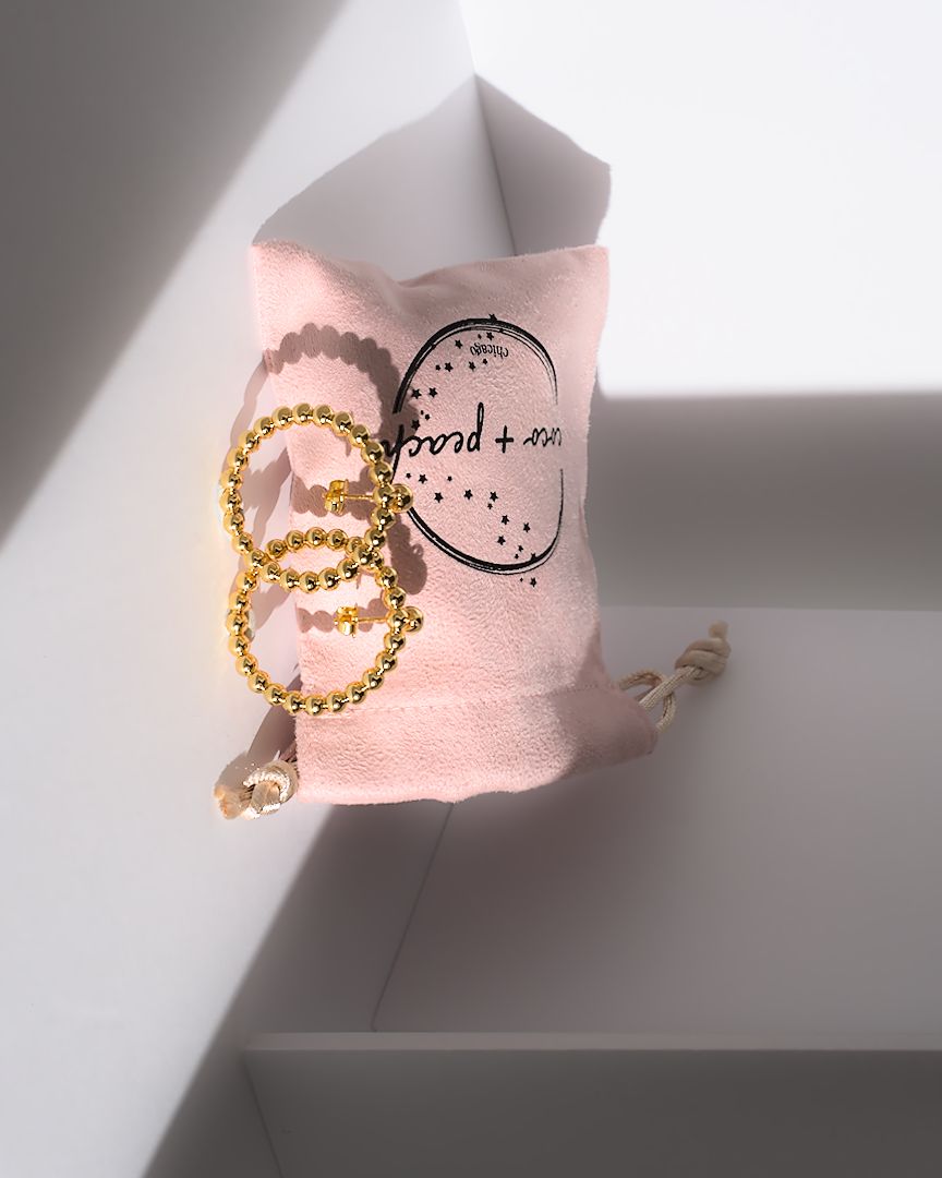 Pink fabric pouch with black text and a circle design, two gold hoop earrings, and a beige cord, on a white surface.