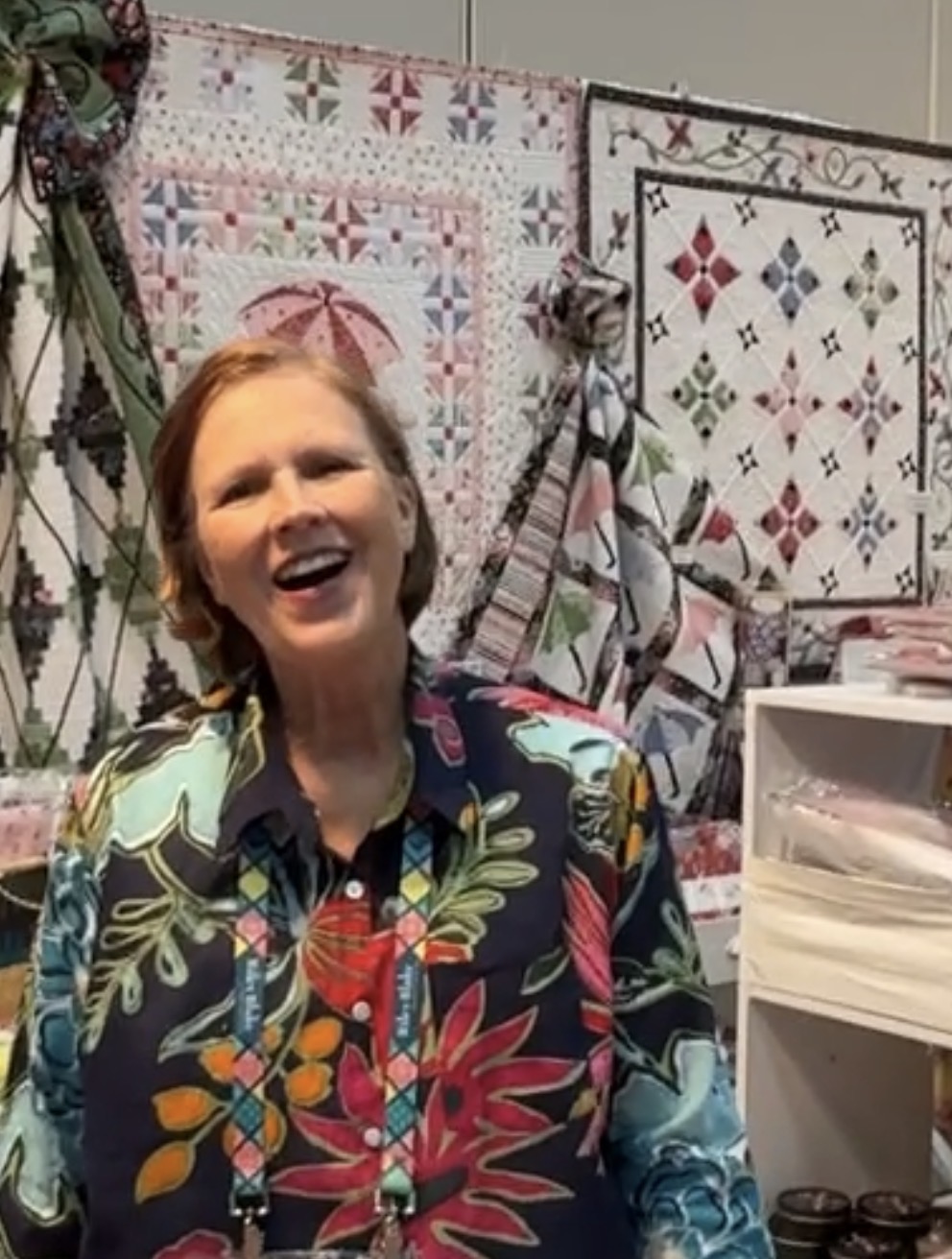 Woman with short hair smiling, wearing a colorful floral shirt, in front of quilted wall hangings.