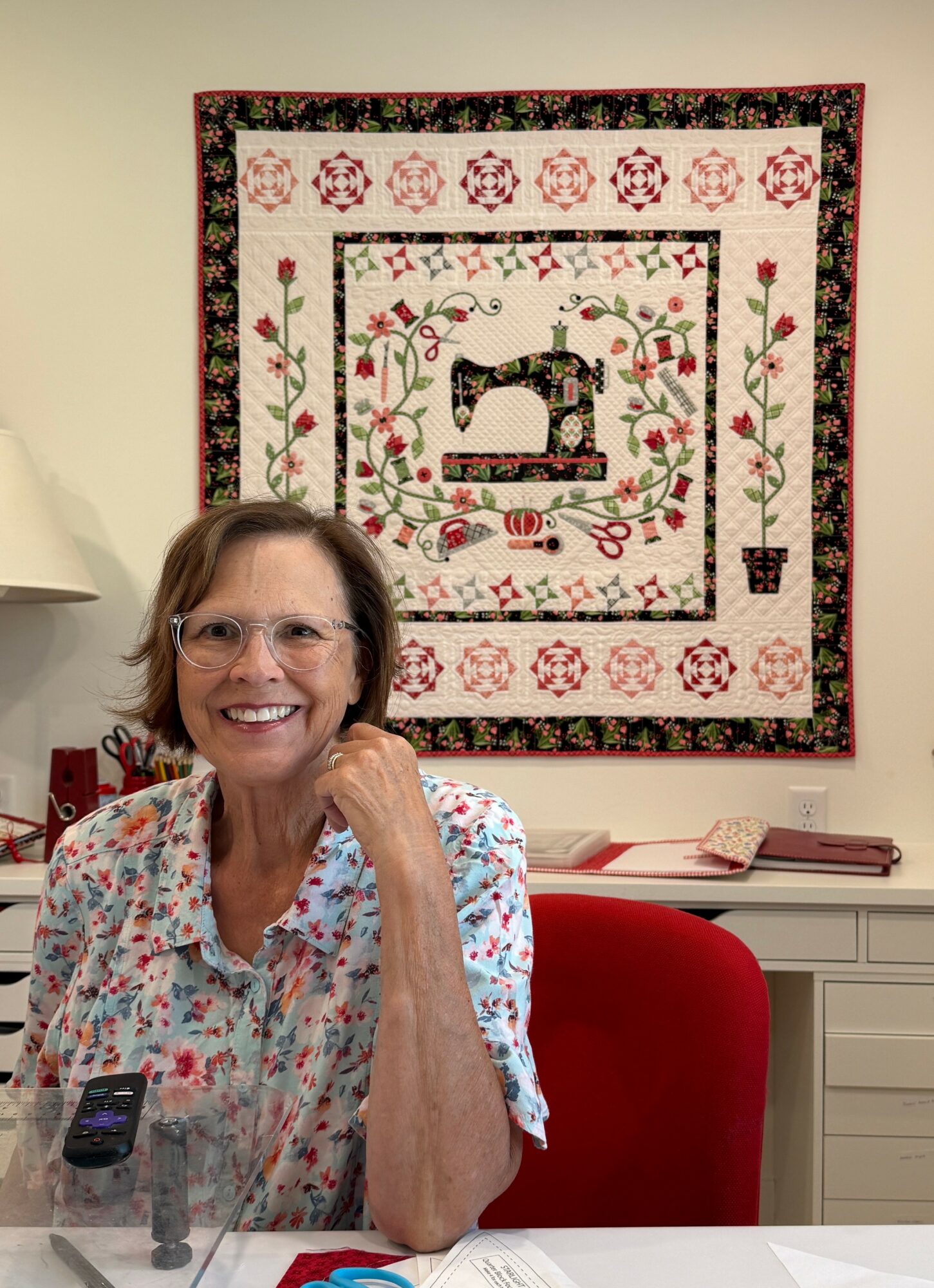 Smiling woman with glasses sitting at a table in front of a quilted wall hanging, wearing a floral shirt, with a red chair.