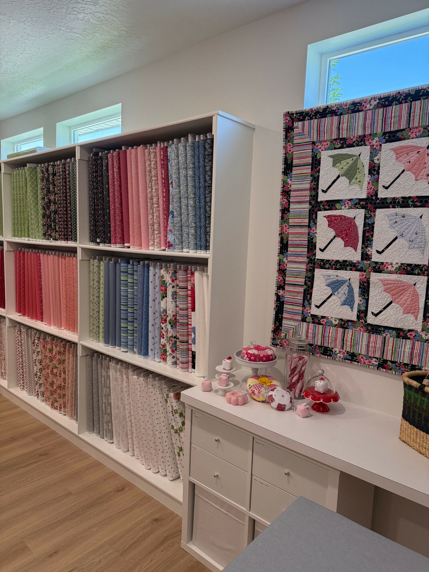 Bookshelves filled with colorful fabric or quilt patterns, a quilt hanging on the wall, and a table with decorative items.