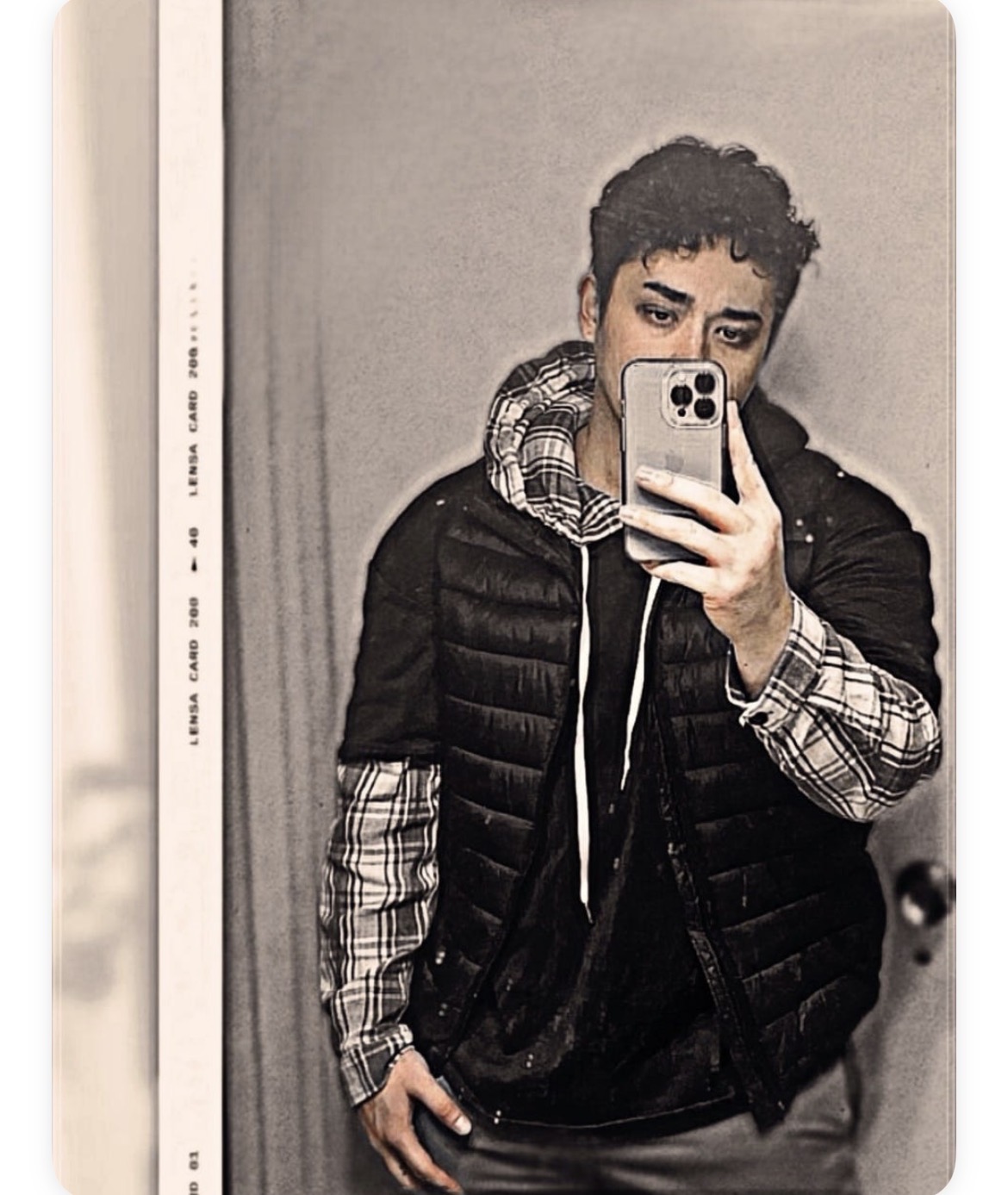 Young man taking a mirror selfie with a smartphone, wearing a black puffer vest and plaid shirt, in a room with plain wall.