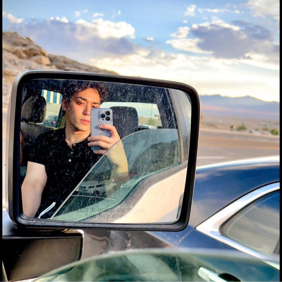 Person taking a selfie in a car's side mirror, with a desert landscape and sky in the background.