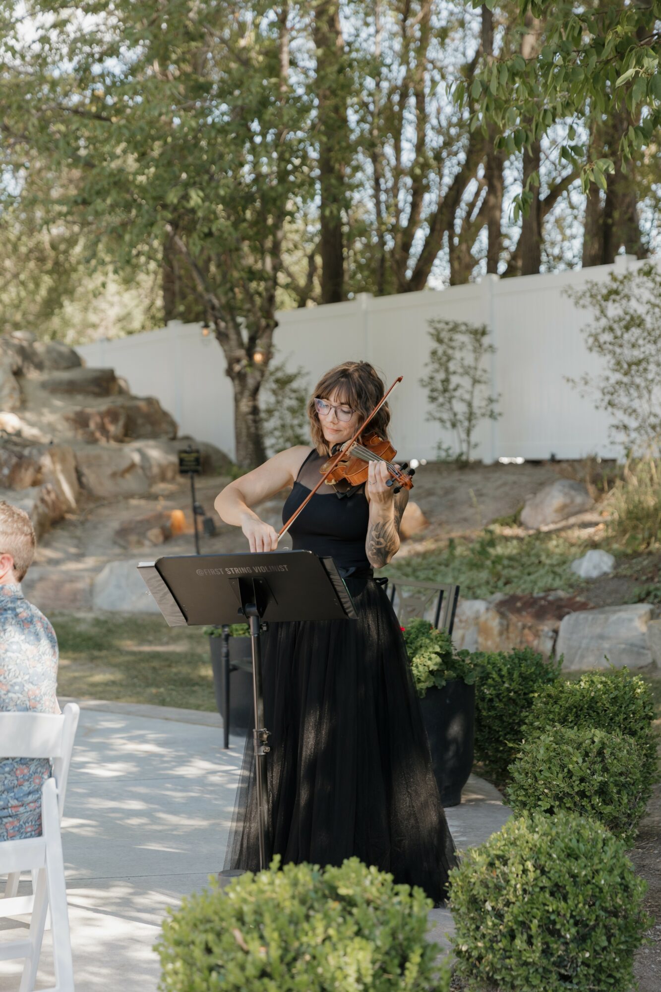 Woman playing violin outdoors near trees and a white wall, with a music stand and greenery around.