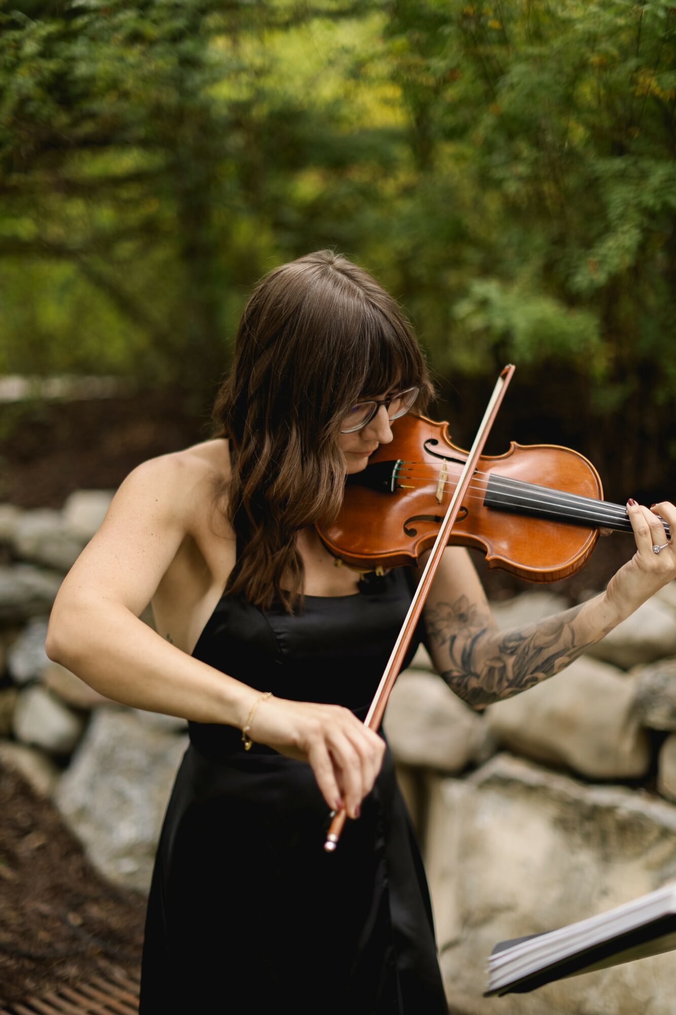 Woman playing violin outdoors with trees and rocks in background, wearing glasses and a black dress.