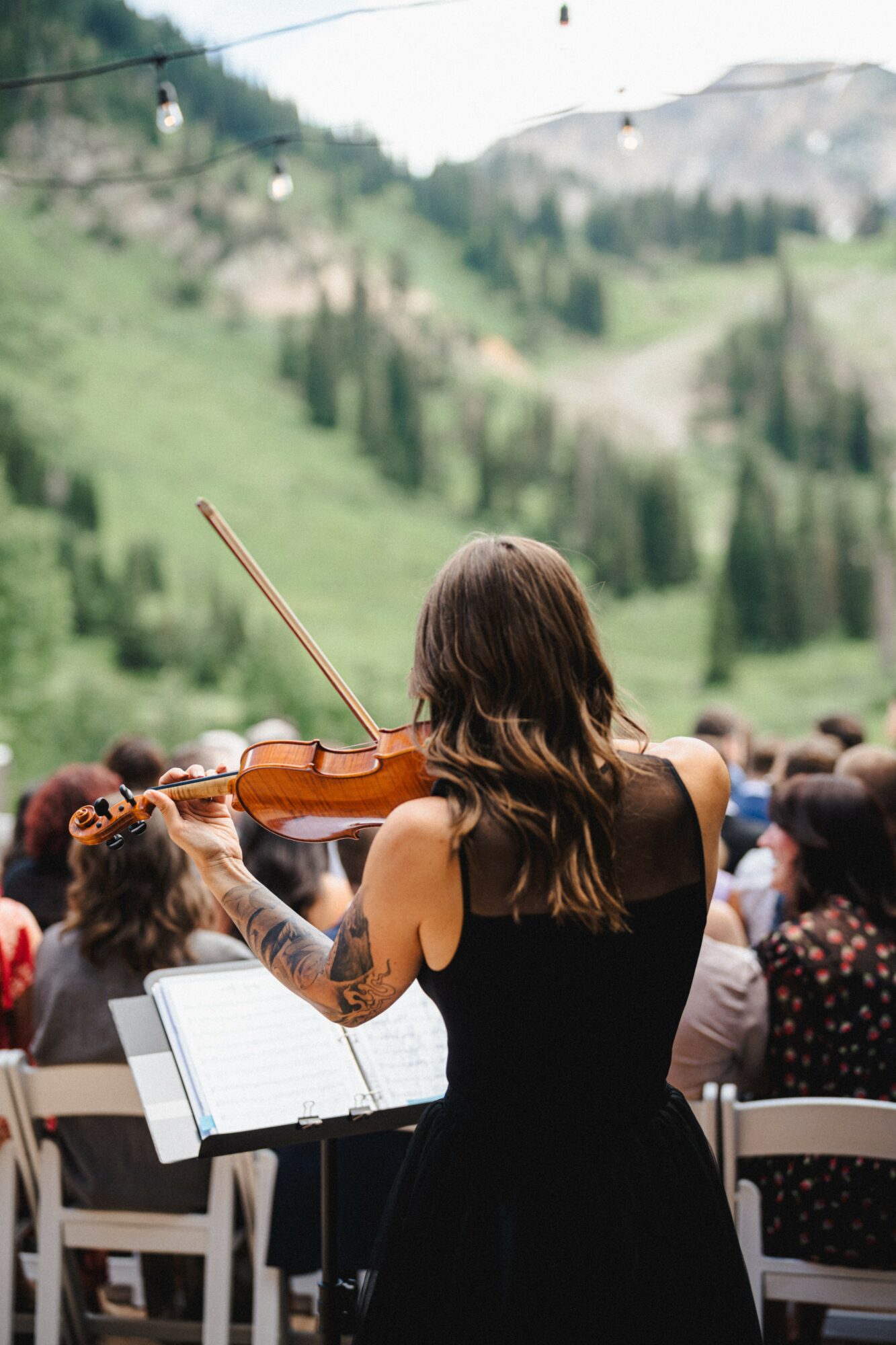 A woman with long hair playing a violin outdoors during daytime, with a mountain landscape and audience in the background.