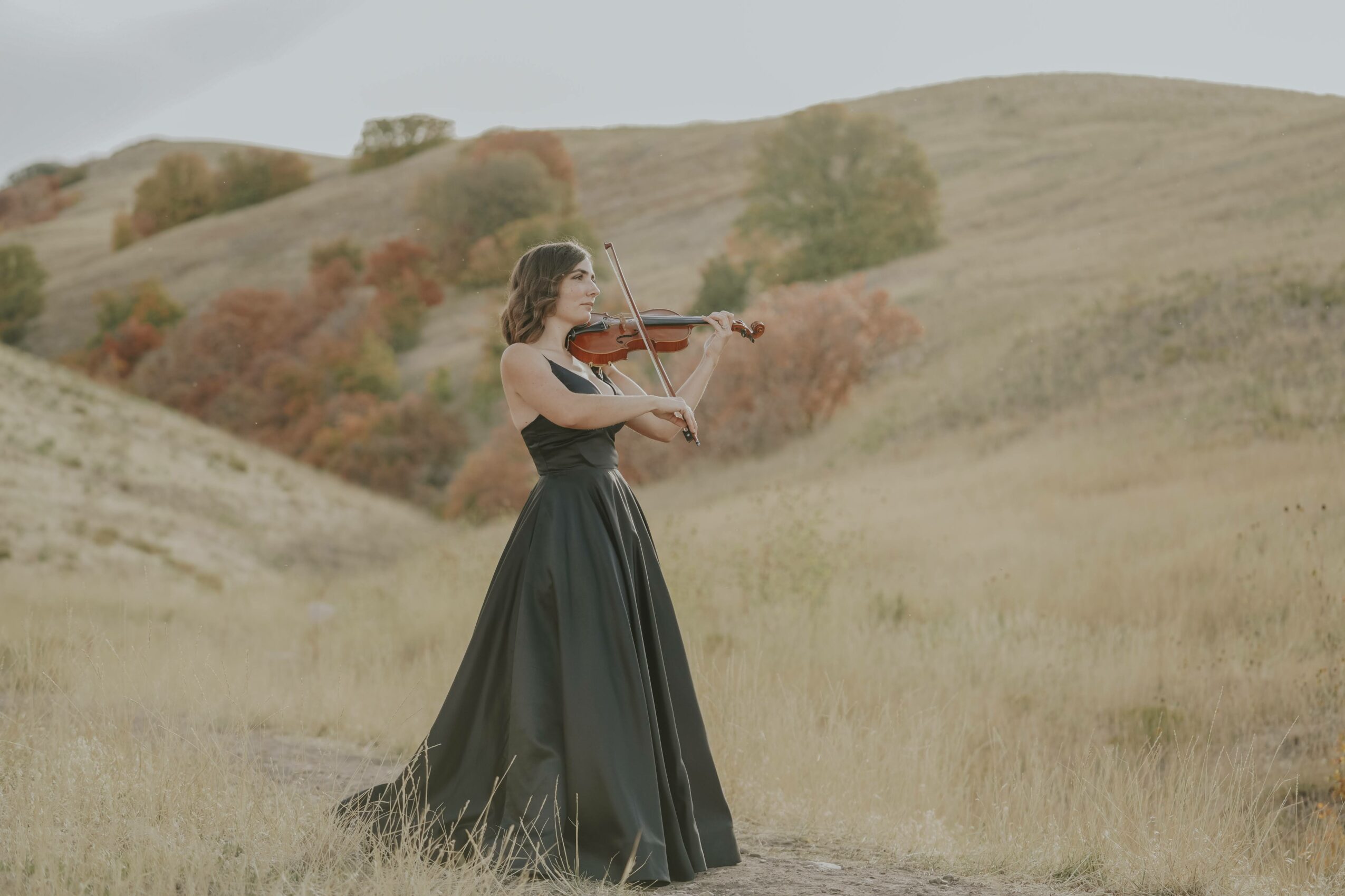Woman in a long black dress playing violin outdoors in a grassy field with rolling hills and trees in the background.