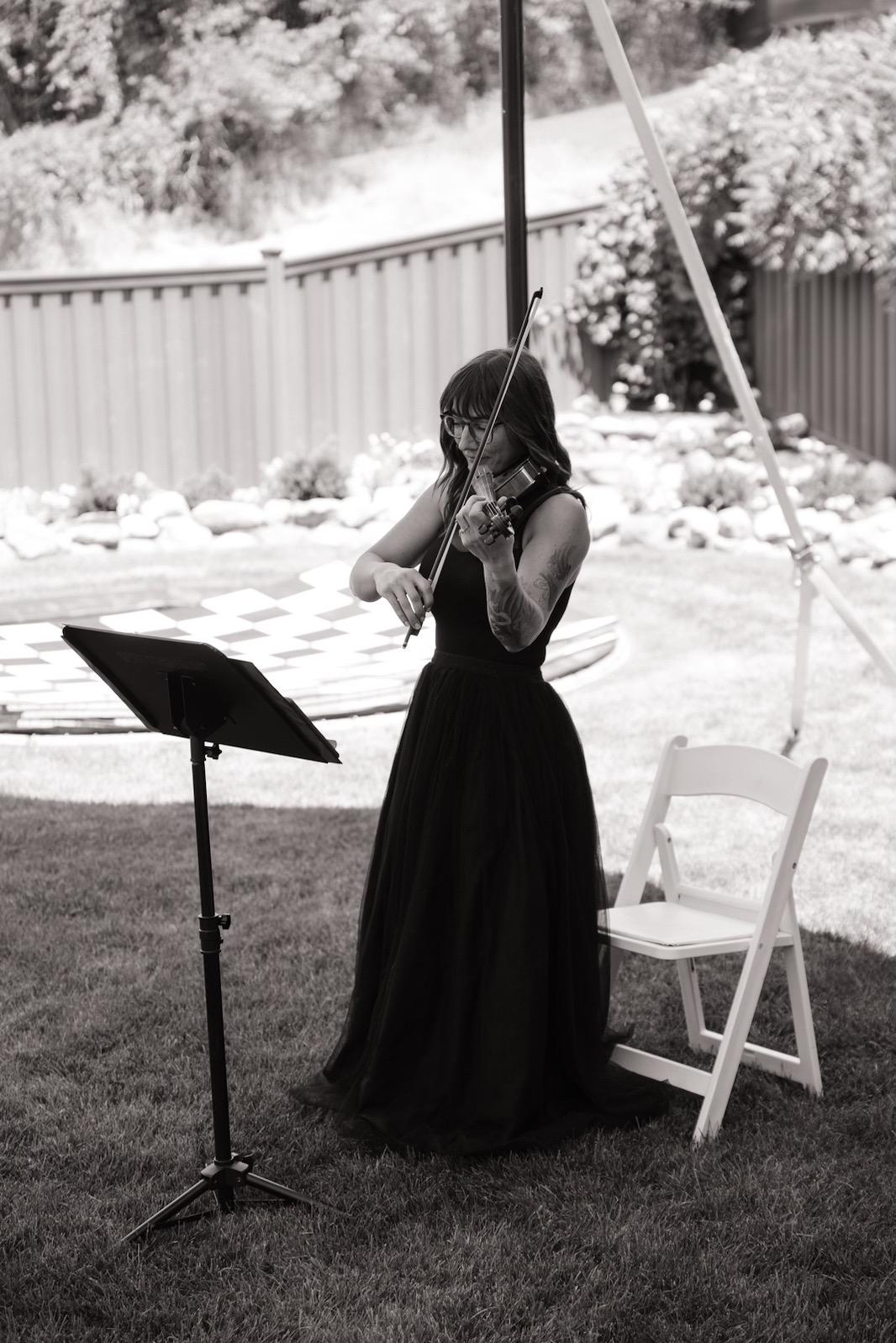 Woman in a long dress playing a violin outdoors with a music stand and a chair nearby, in a garden setting.