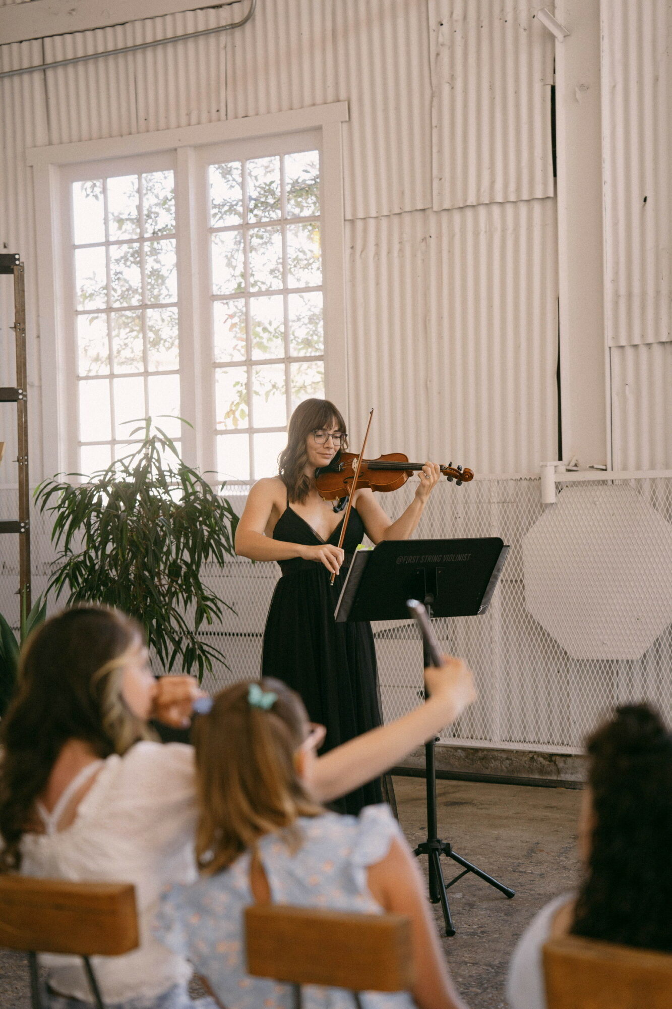 A woman in a black dress plays the violin for children seated in a room with large windows and white walls.