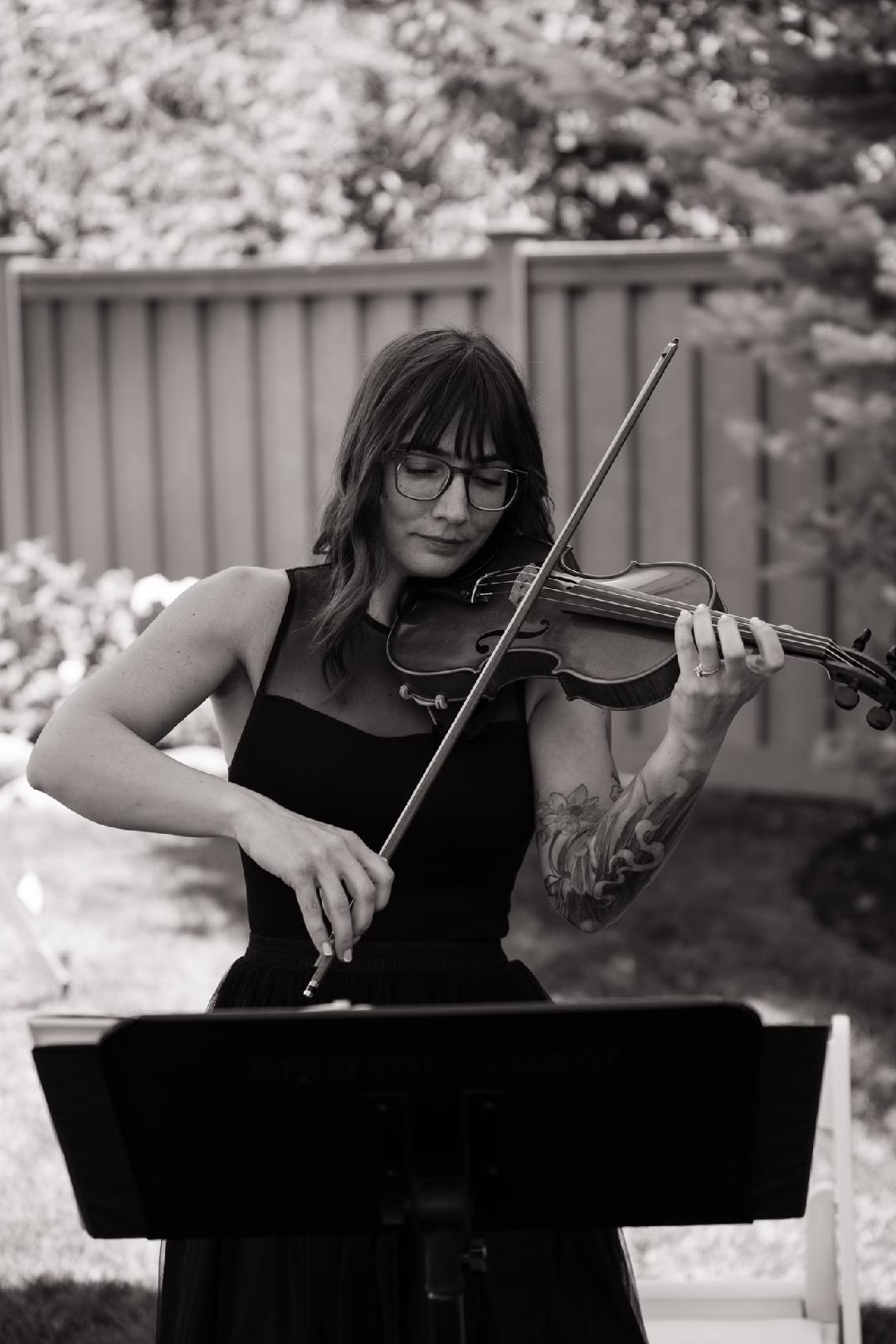 Woman playing violin outdoors, wearing glasses and a sleeveless top, with a music stand in front, background of trees and fence.