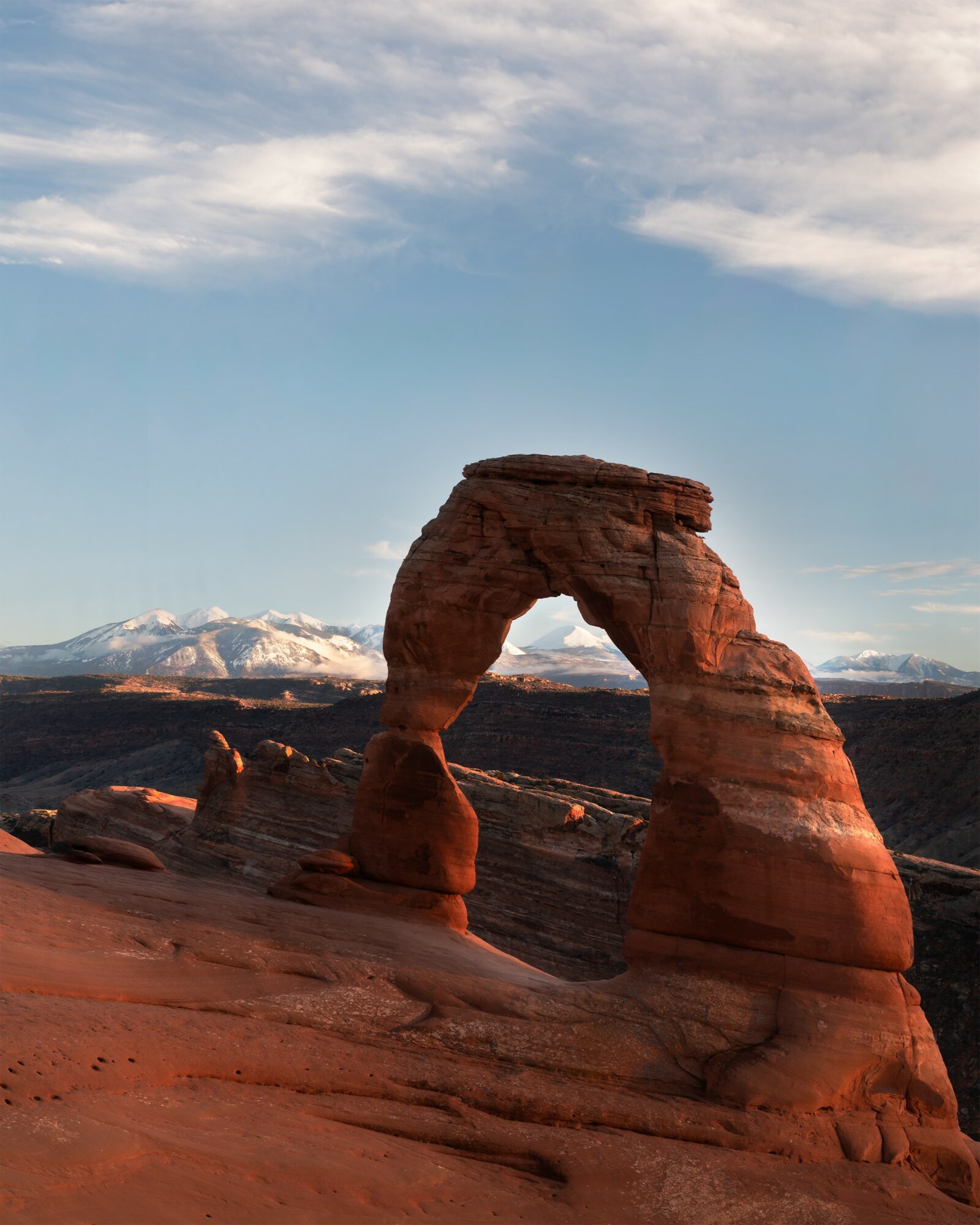 Natural rock formation with an arch in a desert landscape, snow-capped mountains in the background, clear sky.
