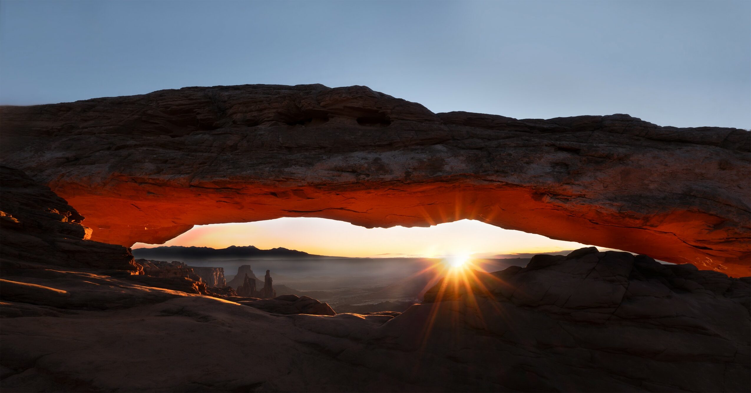 Sunrise or sunset viewed through a natural rock arch with a bright sky and landscape in the background.