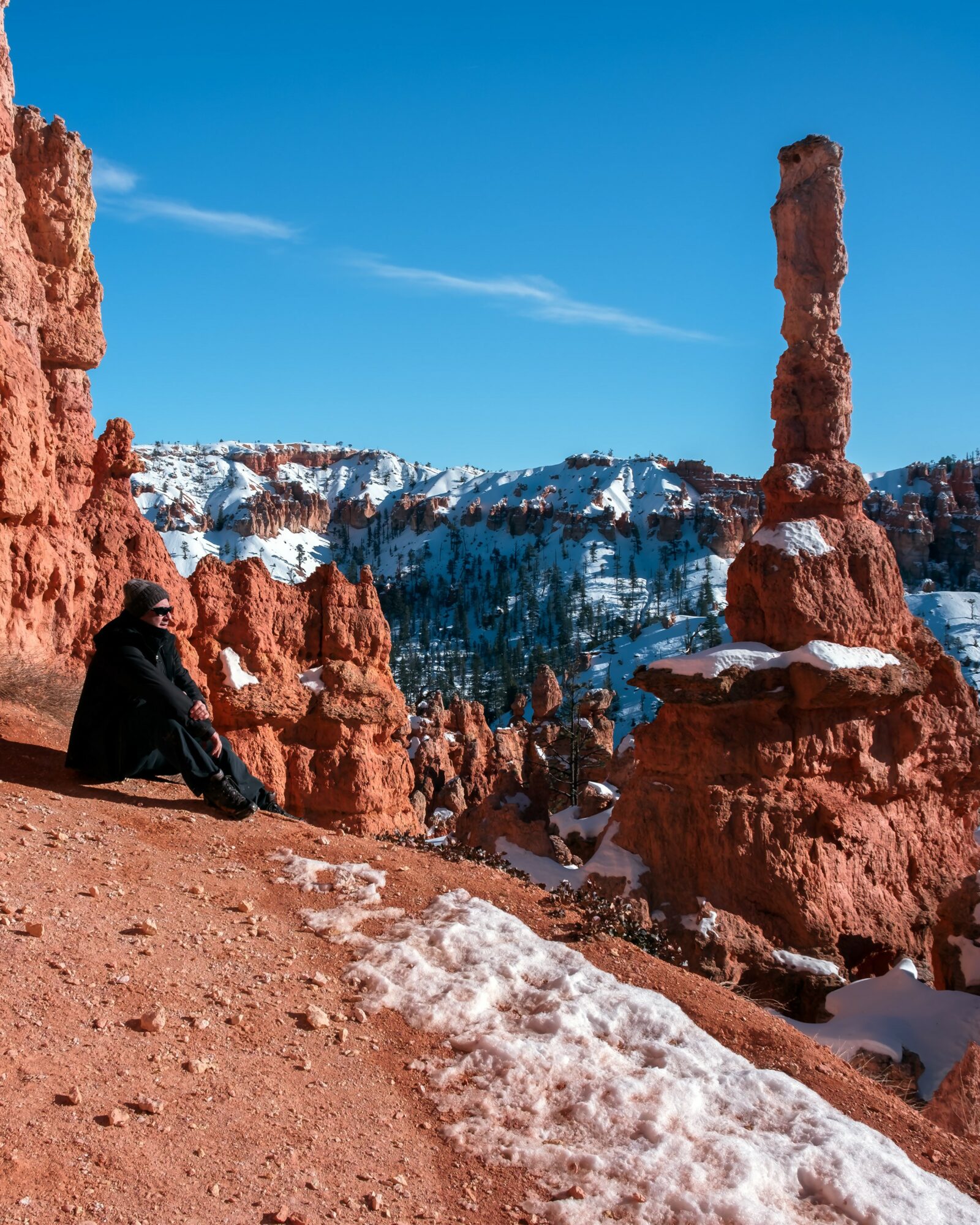Person sitting on reddish rock formation with snow, tall rock spire and snowy mountains in background, clear blue sky.