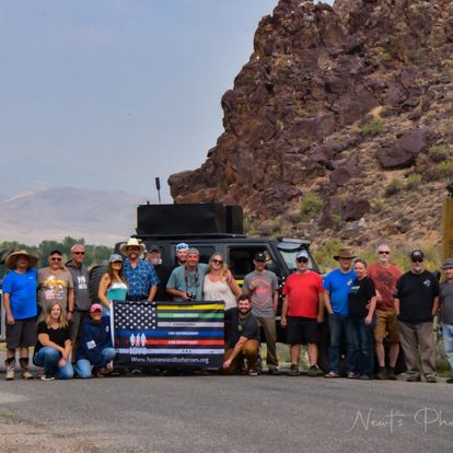 Group of people standing outdoors near a rocky hill, holding a flag with a blue and yellow stripe and a black and white design, in front of a black vehicle.