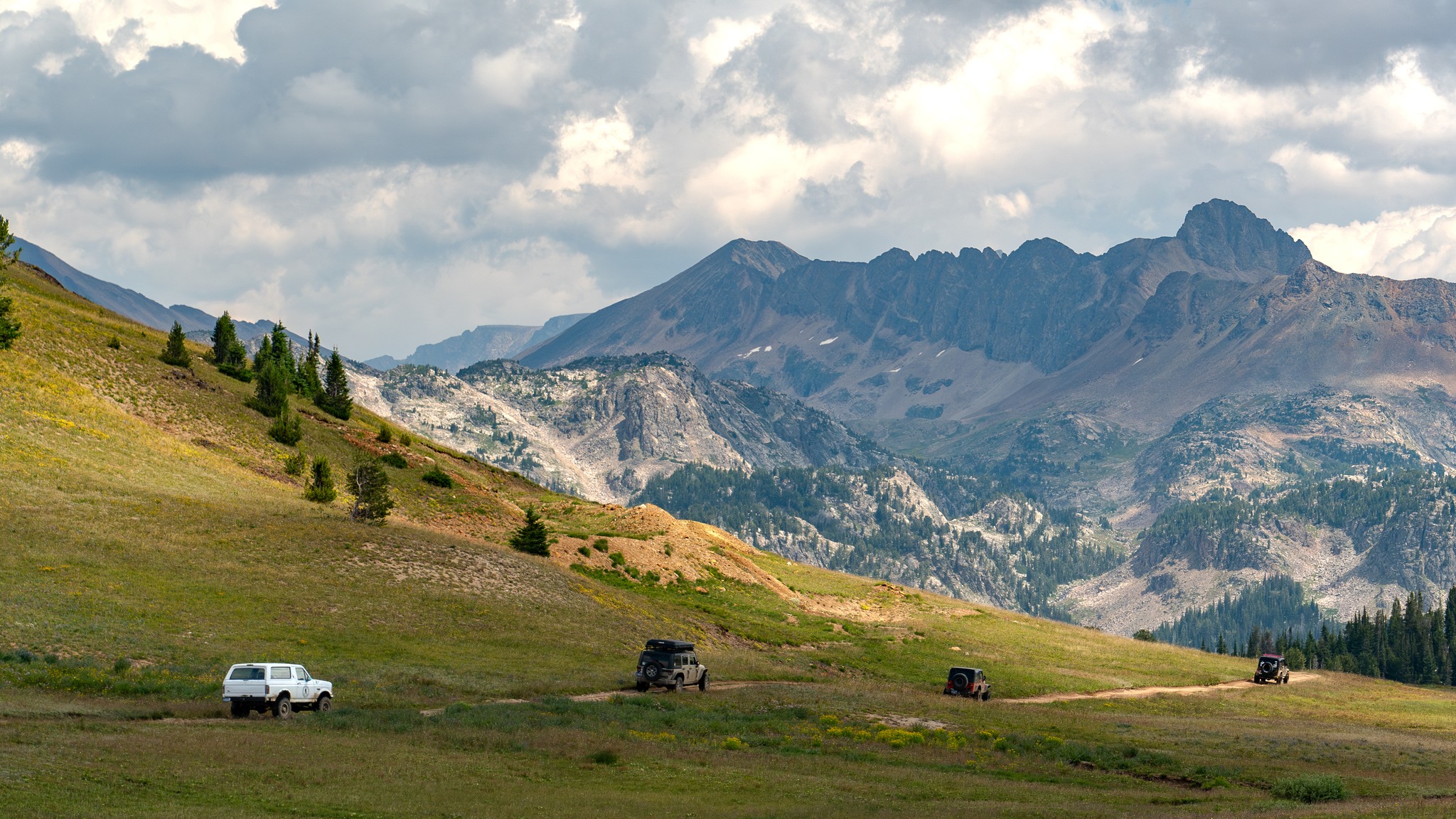 Landscape with mountains, grassy hills, and a few vehicles on a road, under a partly cloudy sky.