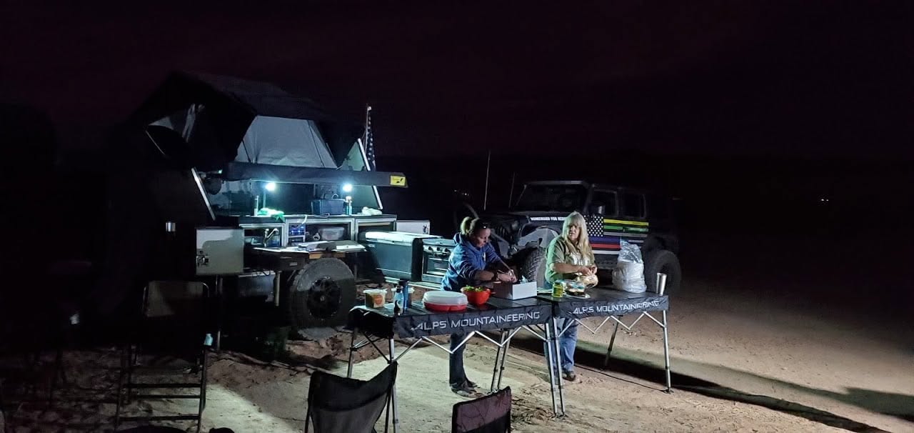 Night scene with a small outdoor setup, people, and equipment illuminated by a light, on a dark background.