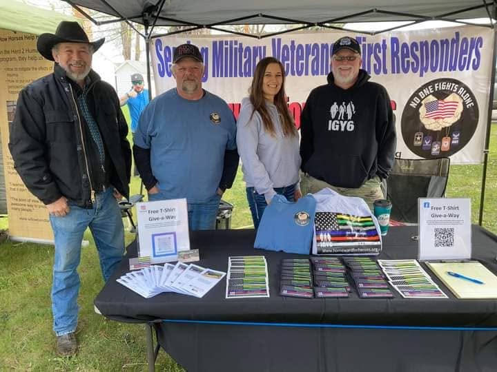 Four people stand behind a table with informational materials and merchandise at an outdoor event, with a banner in the background.