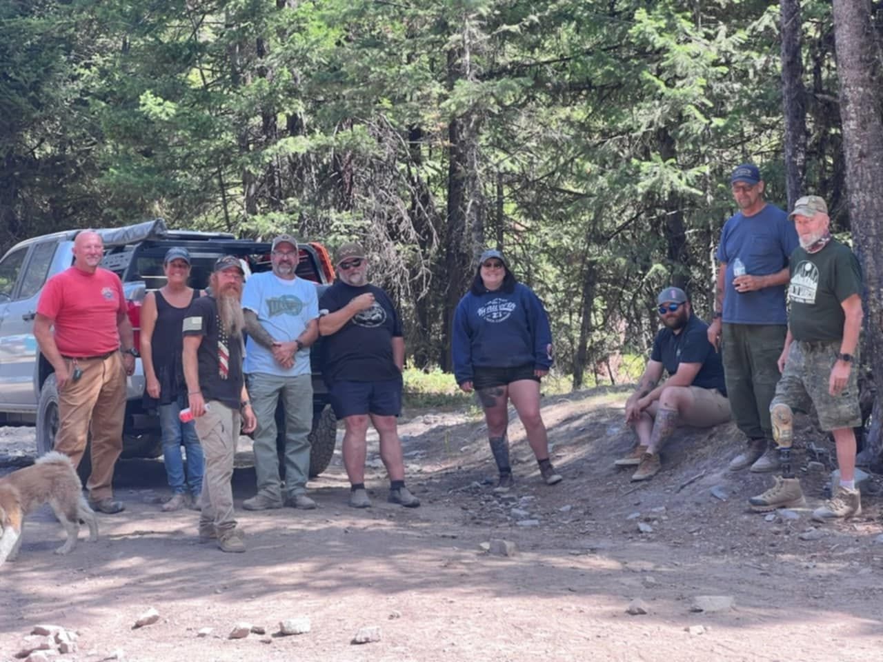 Group of people standing outdoors in a forested area, some with dogs, near a vehicle, with trees in the background.