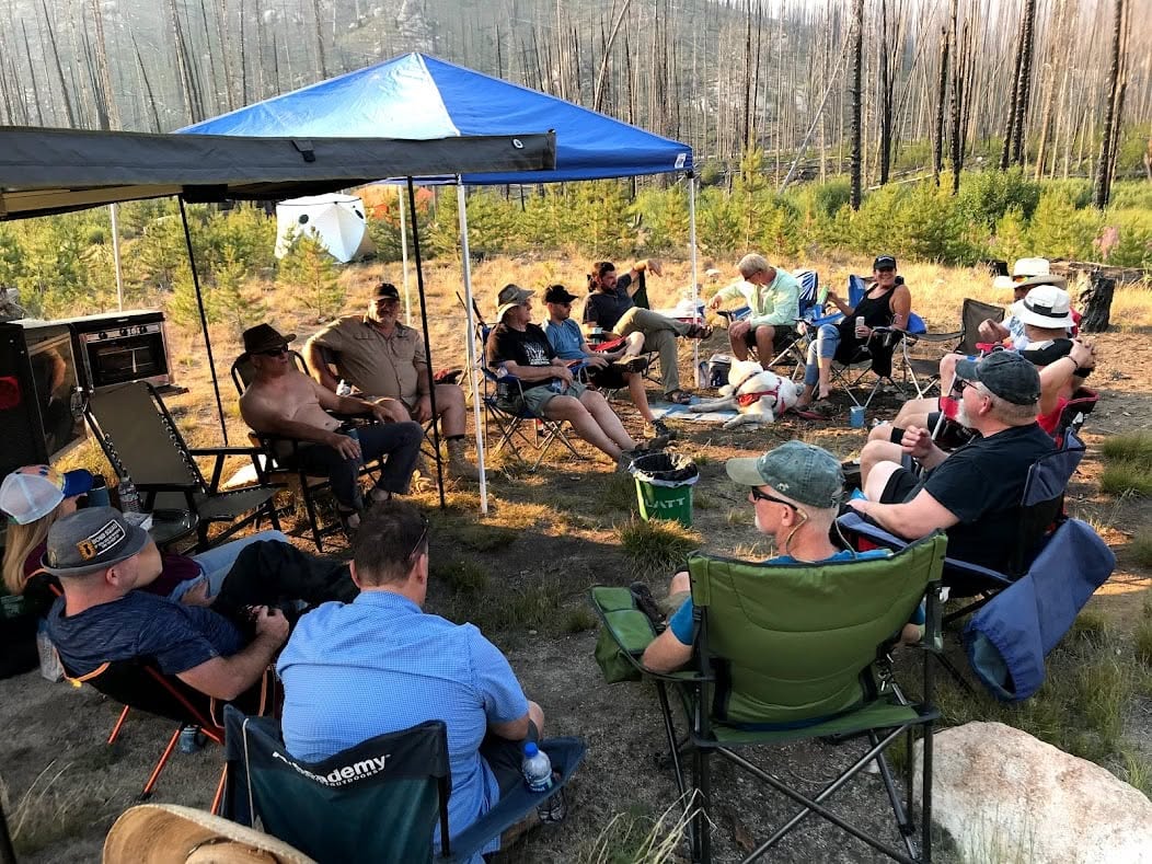 Group of people sitting outdoors under a blue canopy in a forested area, some wearing hats, engaging in conversation.