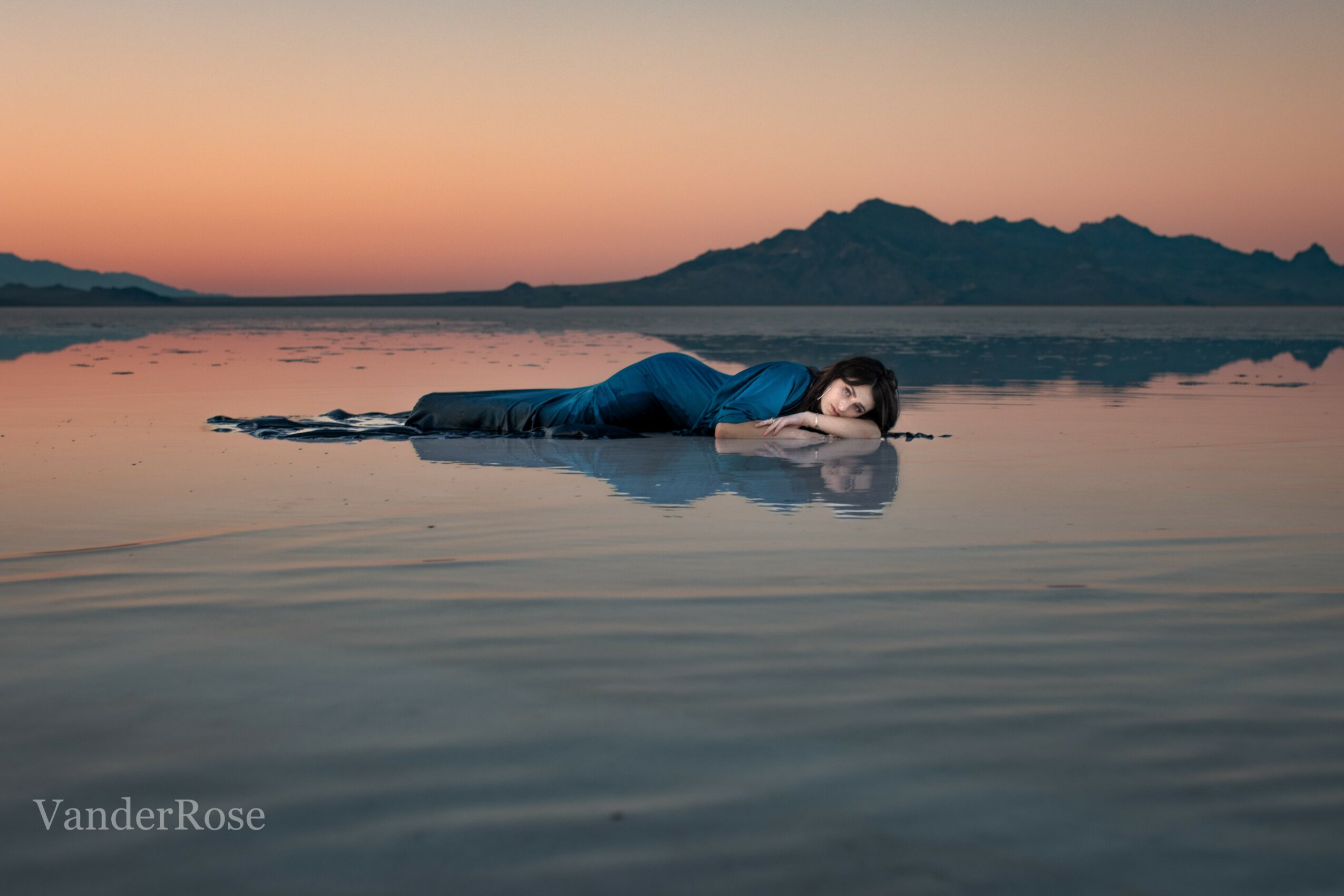 Person lying on water surface during sunset with mountains in background, resting head on hands, calm scene.