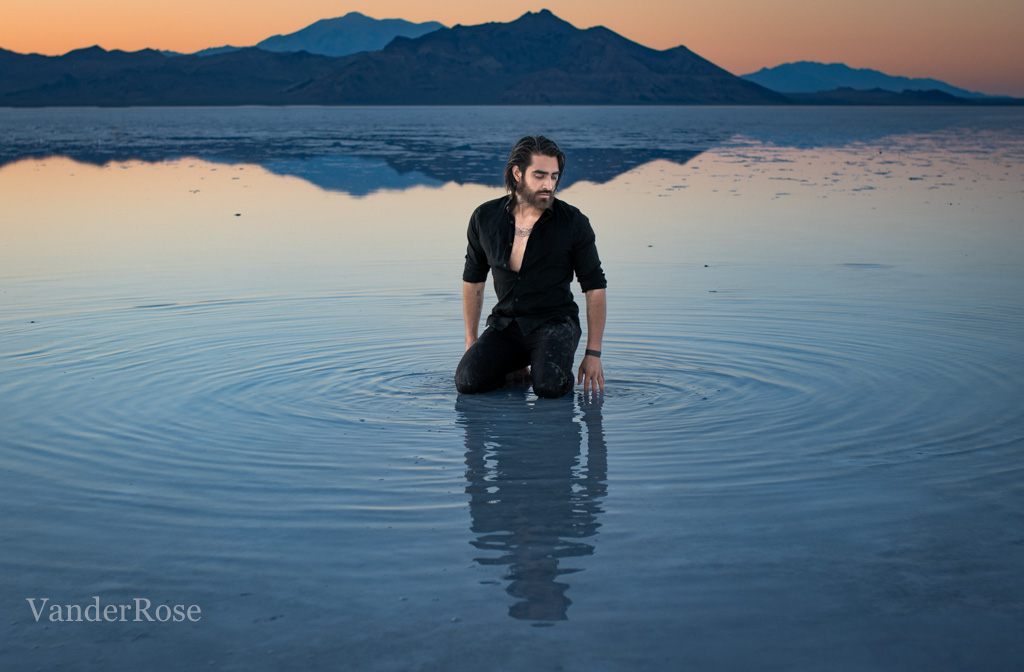 Person kneeling in water with mountains in background during sunset, water reflecting sky colors, calm scene.