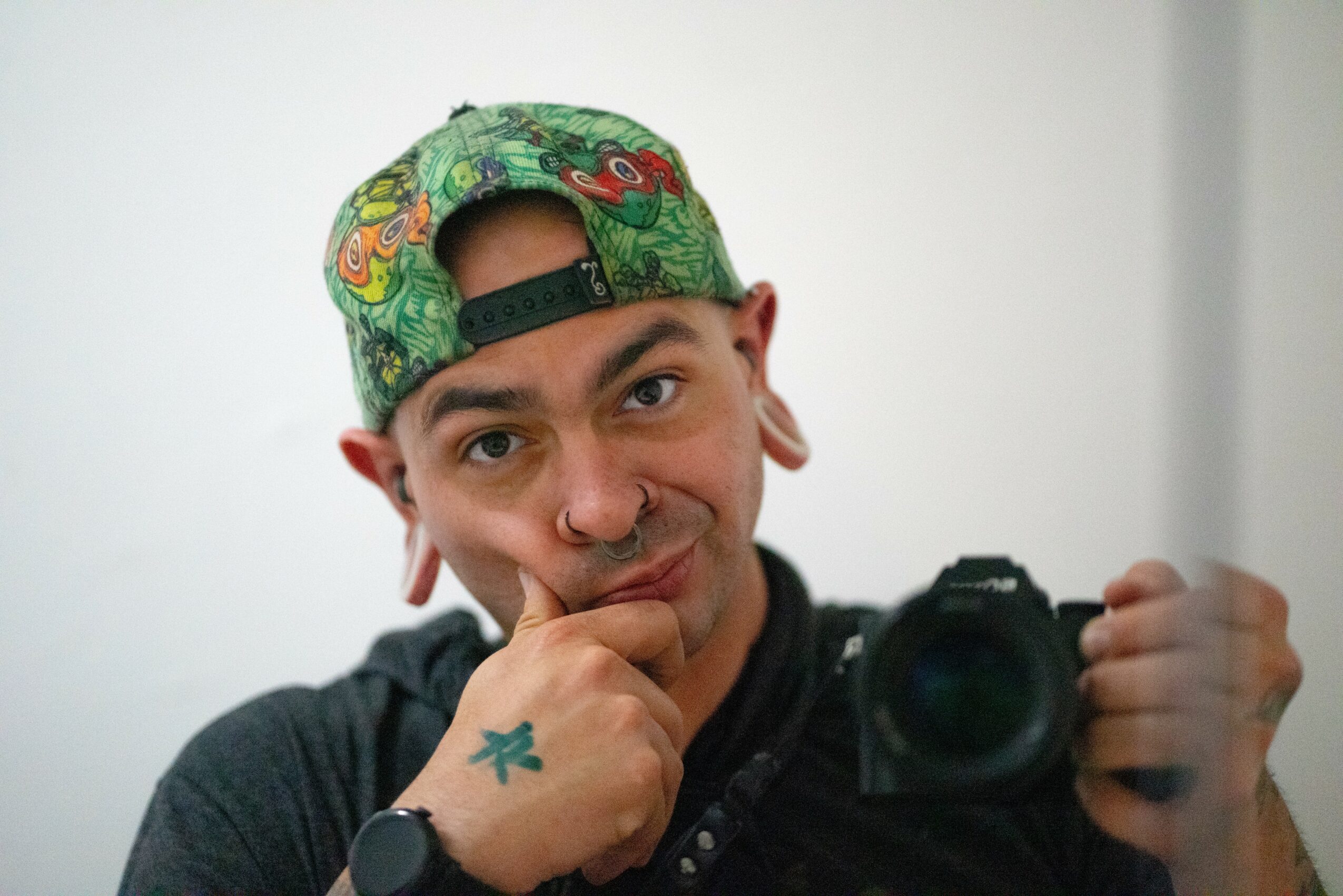Person wearing a colorful cap, holding a camera, with a thoughtful expression, in front of a plain background.