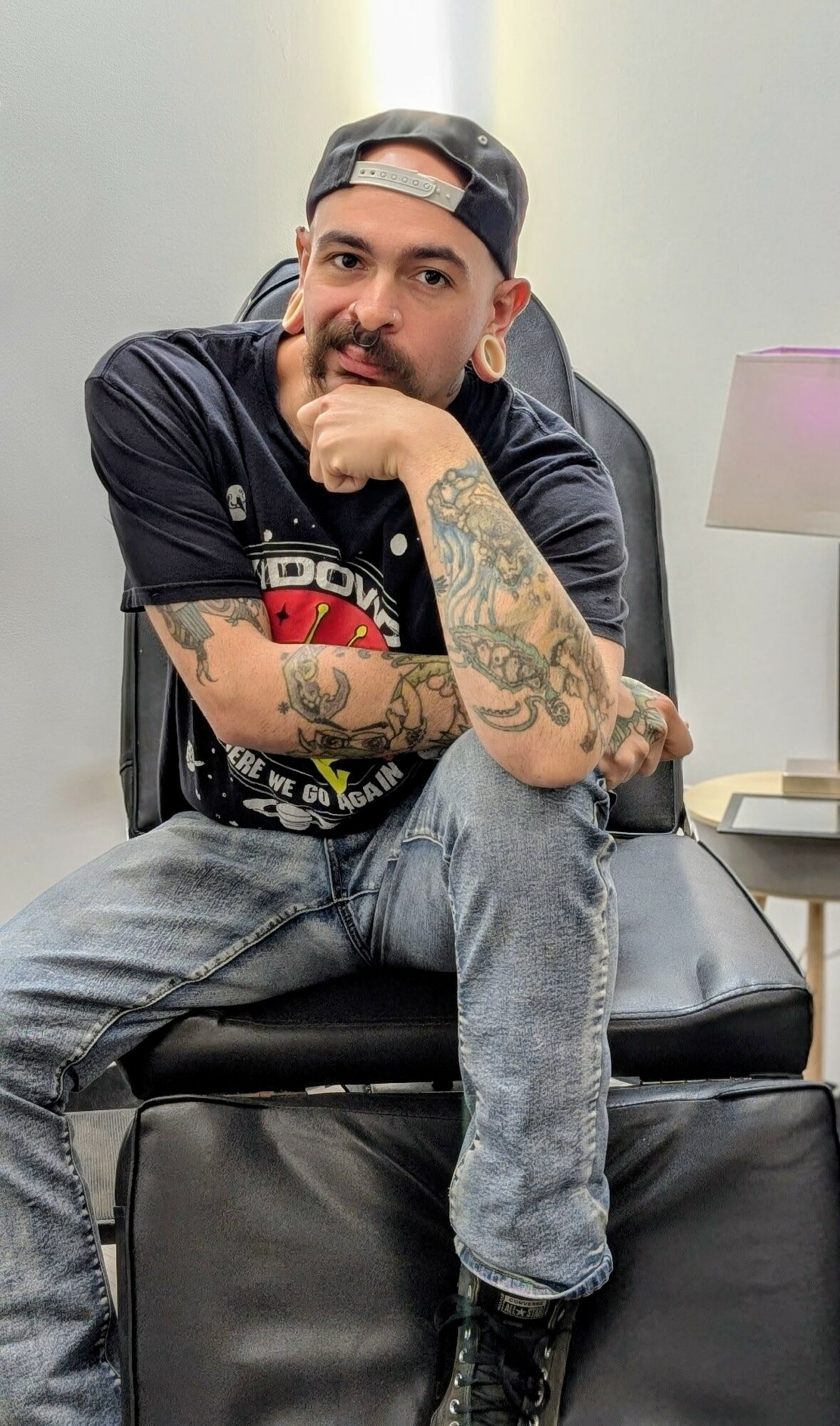 Young man with tattoos, wearing a cap backwards, sitting on a black leather chair, resting his chin on his hand.