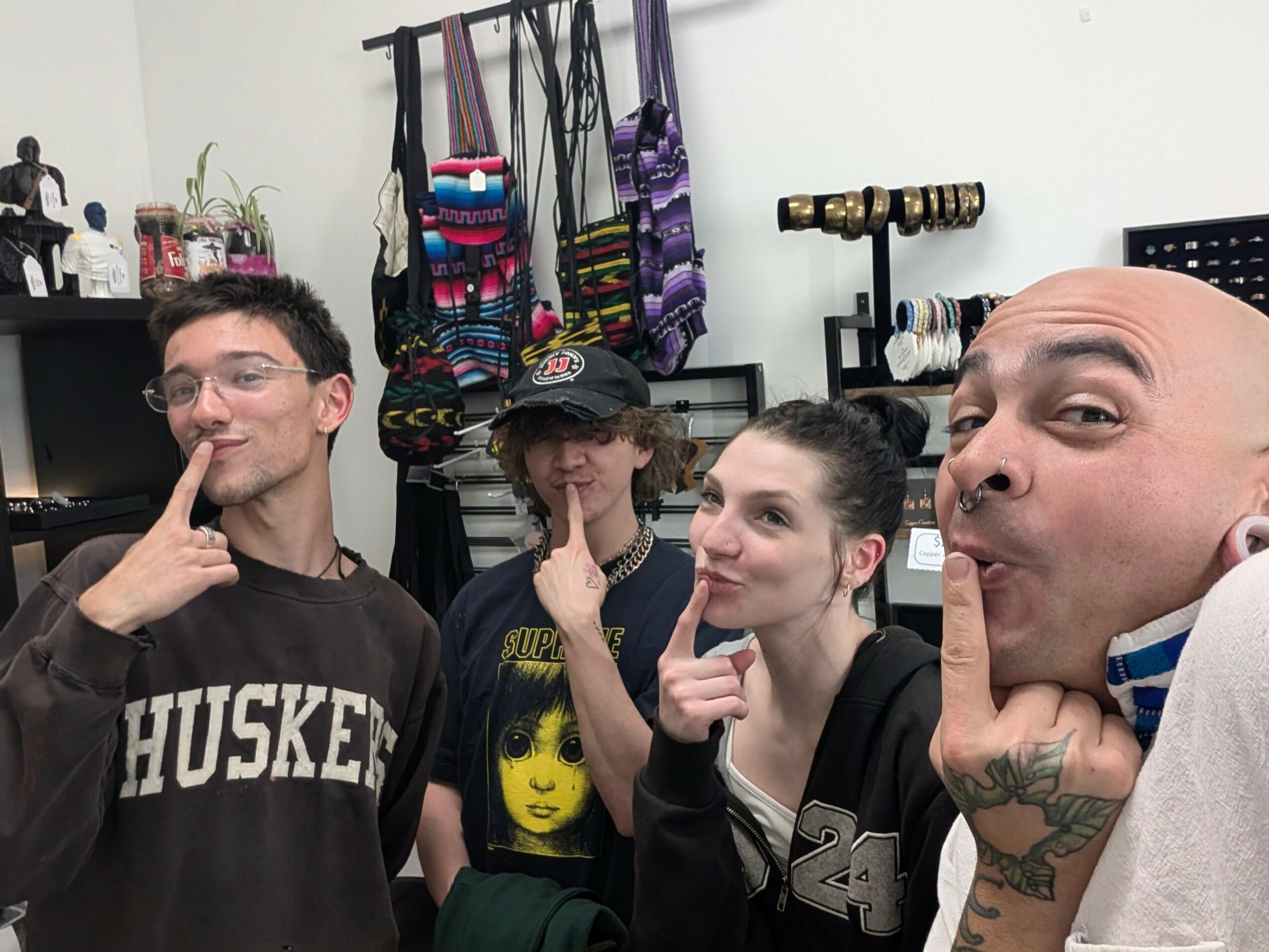 Four people posing indoors, with shelves and hanging bags in the background, all making a finger-to-lips gesture.
