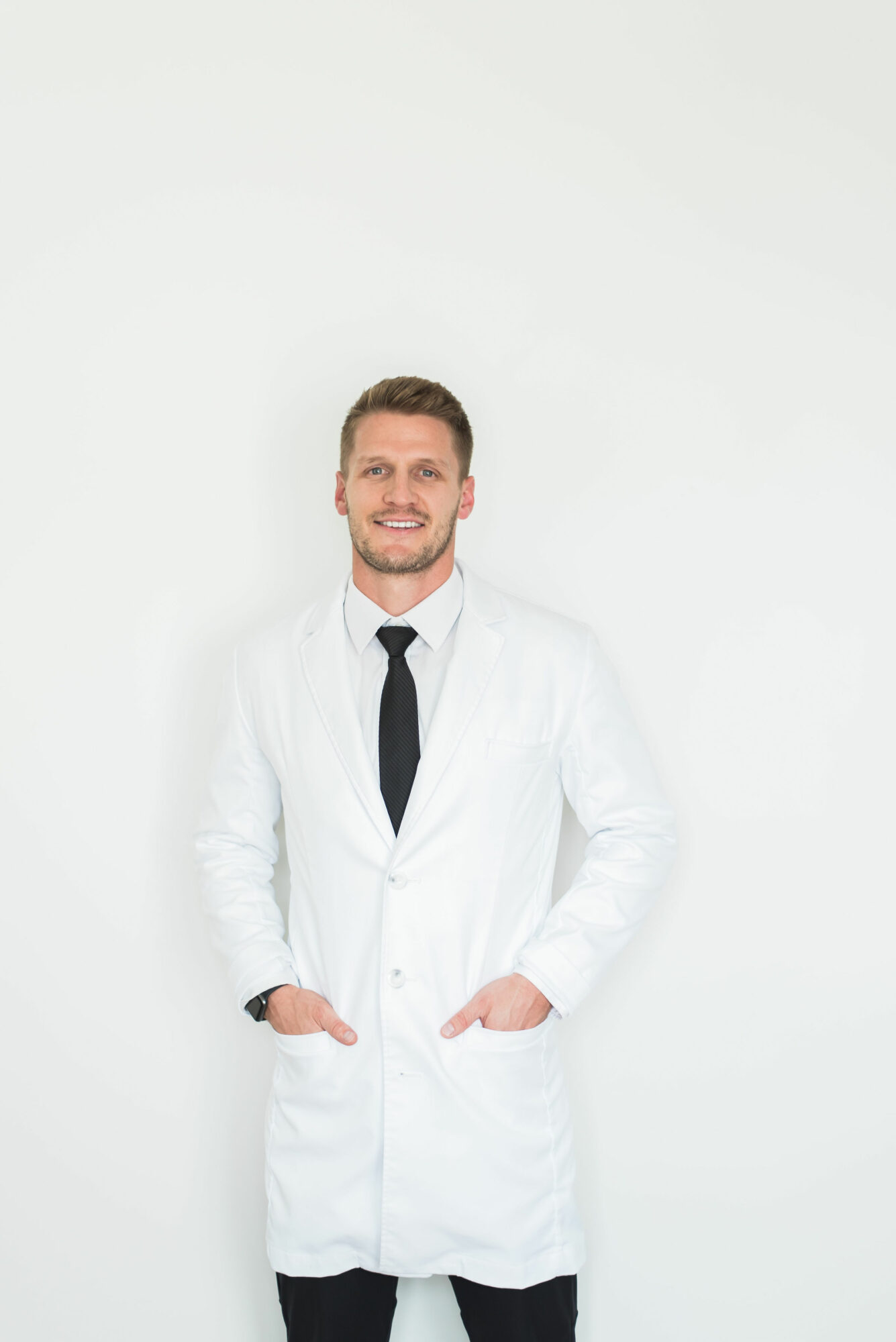 Man in white lab coat and black tie smiling with hands in pockets, standing against a plain white background.