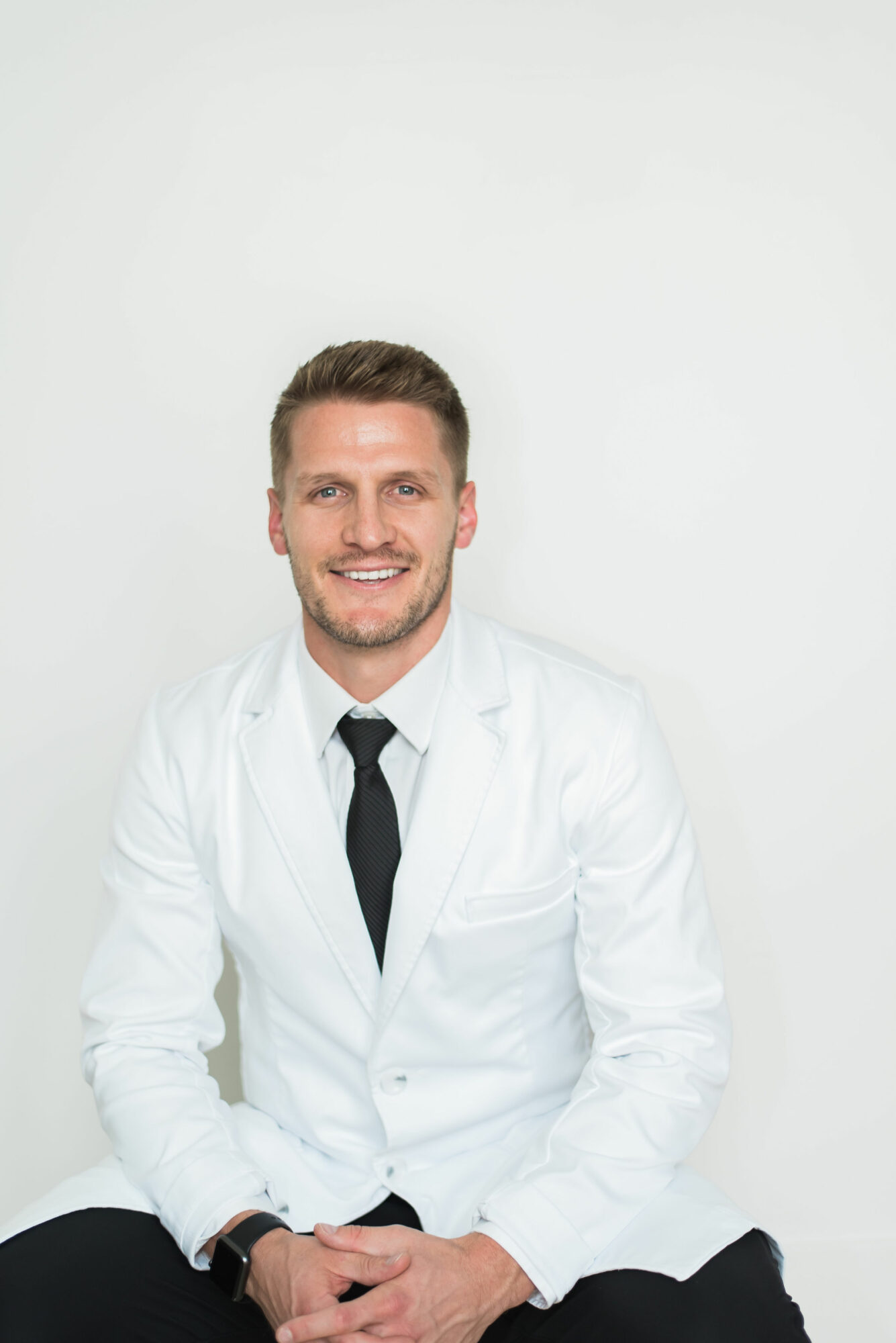 Smiling man in white lab coat and black tie sitting against plain white background.