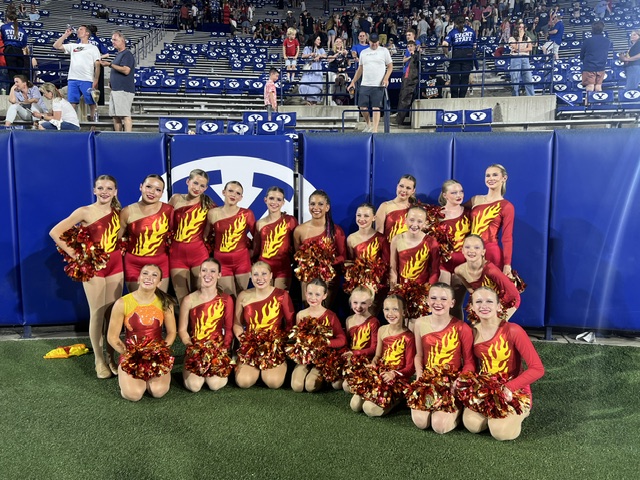 Group of young cheerleaders in red and yellow uniforms posing indoors on a sports field, with spectators in the background.