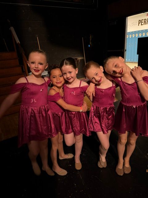 Four young girls in matching pink dresses pose together on stage, smiling and hugging each other.
