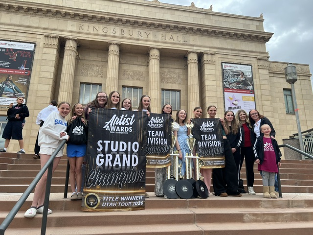 Group of people standing on stairs in front of Kingsbury Hall, holding a large award sign, with a building facade behind them.