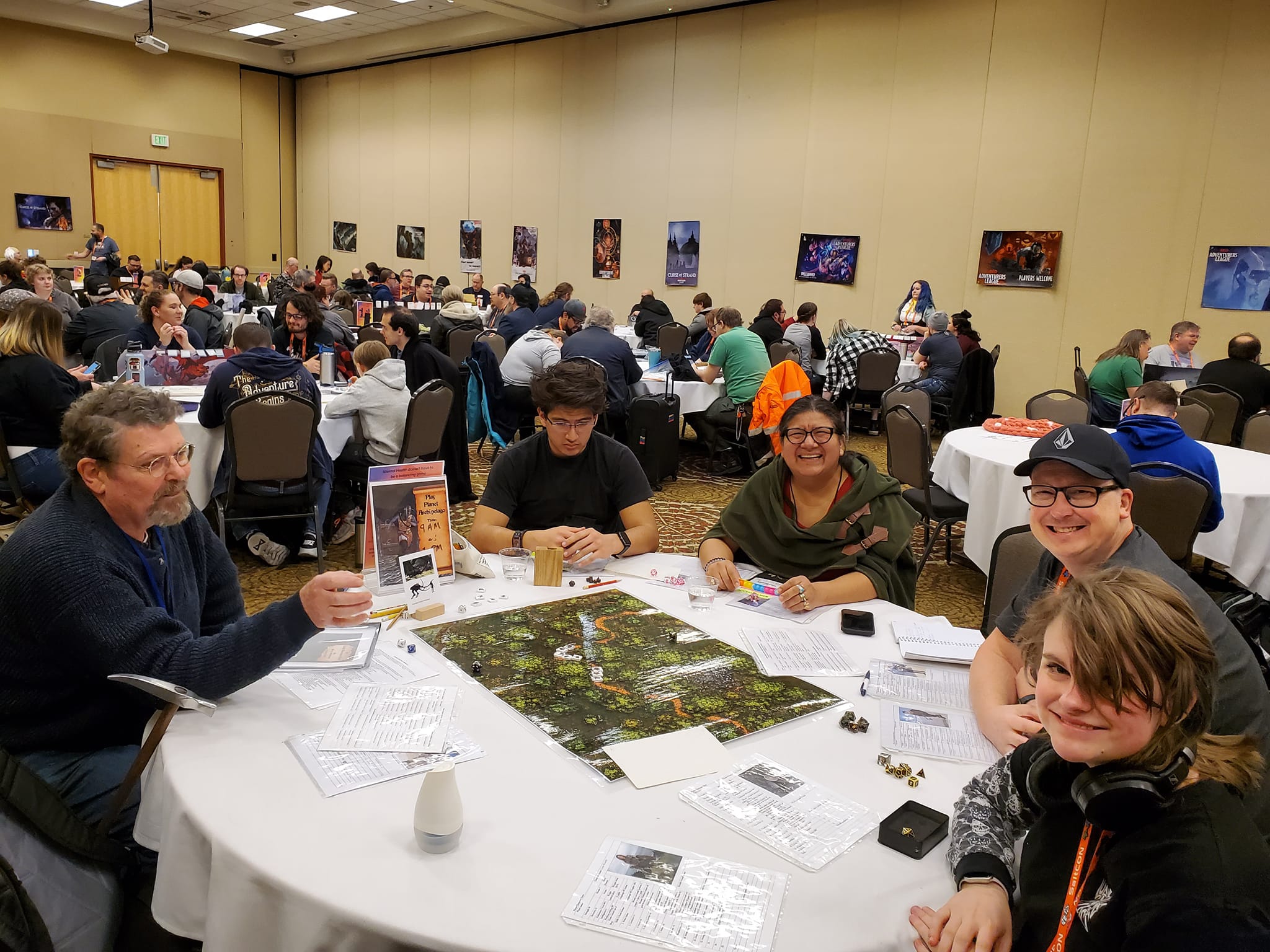 People seated at round tables in a large room, playing a tabletop game with a map, surrounded by posters on the walls.