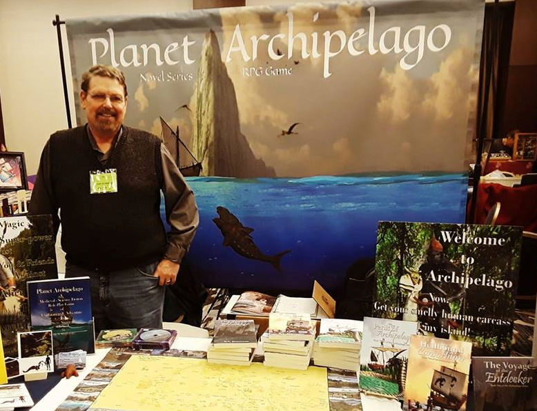 Man standing at a booth with books and a large banner reading 'Planet Archipelago' behind him.