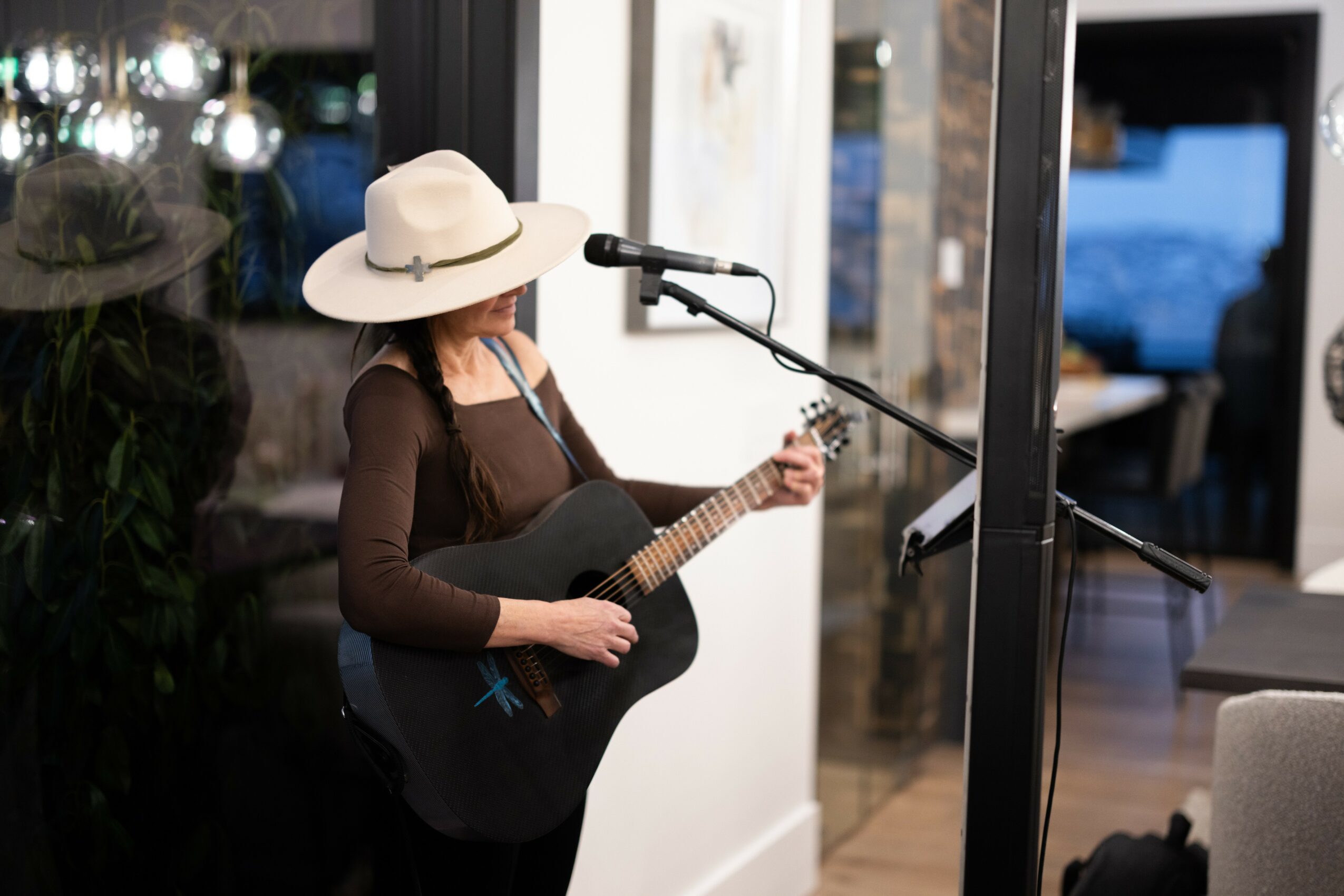 Person wearing a wide-brimmed hat playing guitar and singing into a microphone indoors.