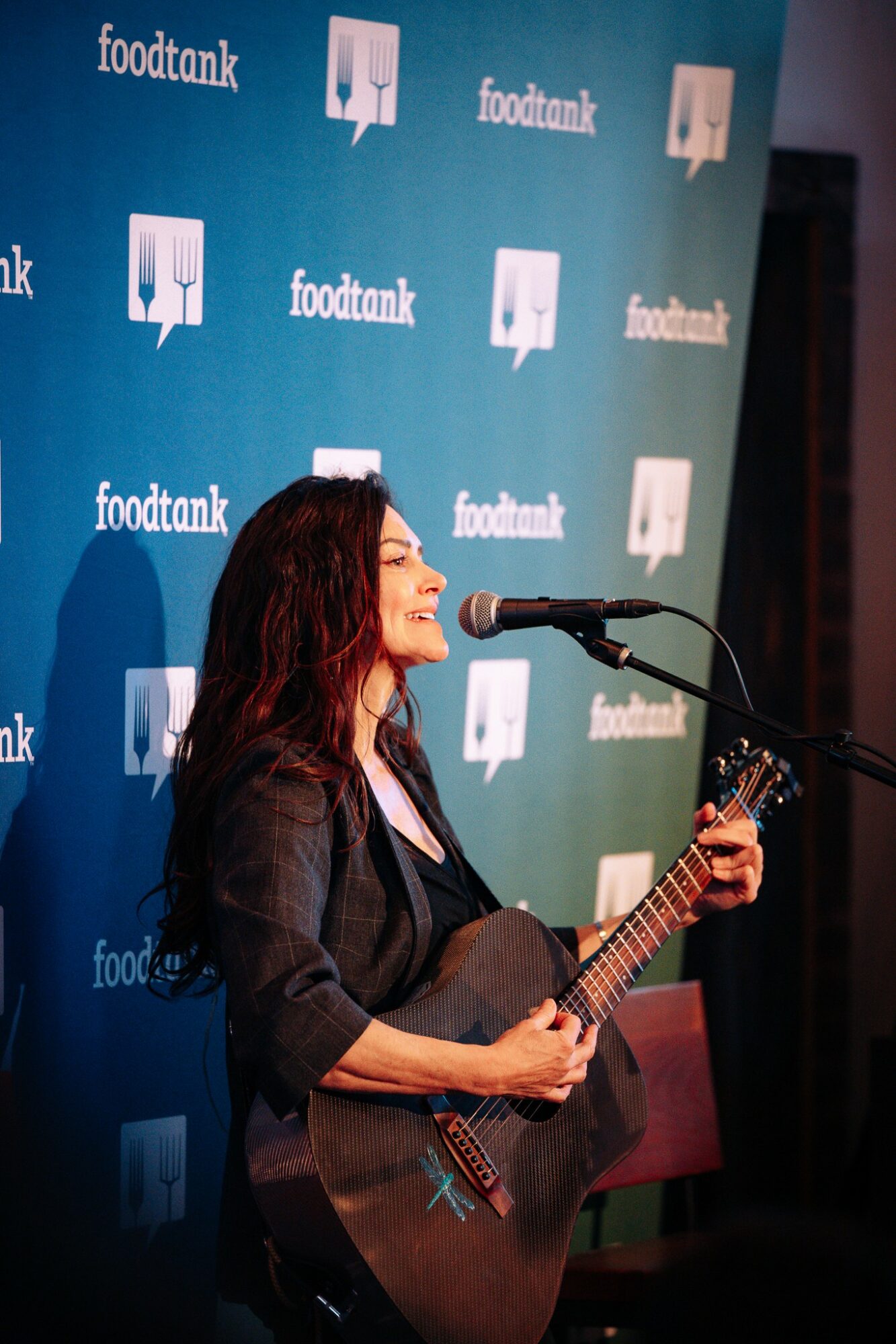 Woman with long dark hair playing guitar and singing into a microphone at an event, with a blue backdrop.